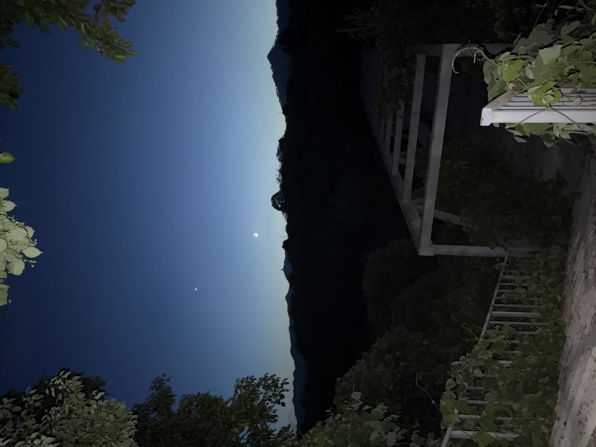 A nighttime exterior view shows a dark landscape with a clear, deep blue sky. The sky features a crescent moon and a bright star. In the foreground, overgrown vines climb a white railing leading down a set of stairs. To the right, a partially constructed wooden pergola or trellis stands. Beyond the immediate foreground, a silhouette of forested hills stretches across the horizon. A lone structure or tower is visible on one of the distant hills. The overall scene suggests a rural or semi-rural setting in the city of Jiangjin, China, likely a residential area or a scenic overlook after dusk.