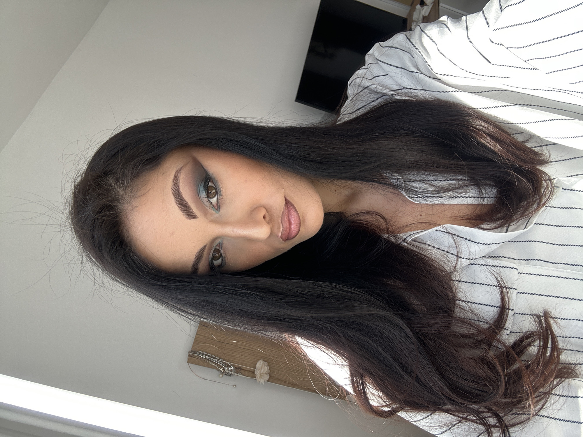 A woman with long, dark hair poses for a close-up, indoor portrait. Her face is tilted, and she is looking directly at the camera. She has a full face of makeup including blue eyeshadow, heavy eyebrow makeup, and nude lipstick. She is wearing a white button-down shirt with thin black pinstripes. A TV is visible in the background, mounted high on a wall. Part of a decorative wooden board with a few silver chains and a small white flower is partially visible in the lower left. The location is Unknown, Unknown.