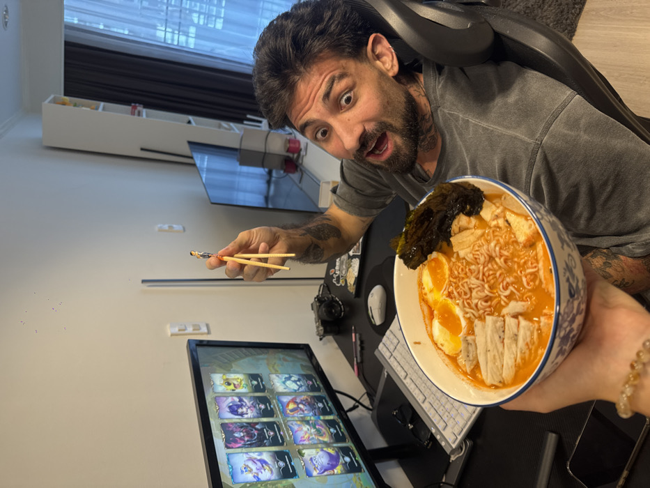 The scene depicts an indoor setting within a modern apartment in Bangkok, Thailand. A male individual, appearing to be in his 30s or 40s, with dark hair, a beard, and extensive visible tattoos on his arms and neck, is seated in a black ergonomic chair. He wears a grey t-shirt and displays an exaggerated expression of surprise or delight, with wide eyes and an open mouth directed towards the camera. His right hand holds a pair of light-colored chopsticks, which are gripping a small, male action figure in a red and blue costume, positioned above a large ramen bowl.
The ramen bowl, held in the foreground by an unseen person's hand (revealing a beaded bracelet on the wrist), contains instant noodles in an orange-red broth, two halved boiled eggs, slices of white protein, and a large piece of nori seaweed. To the man's left, a black computer desk is visible, featuring a monitor displaying a video game interface with eight distinct character portraits, a white mechanical keyboard, a grey computer mouse, and a black digital camera. The background consists of white walls, a tall white shelving unit, and a large window with dark curtains, offering a partial view of exterior high-rise structures, consistent with an urban metropolitan area. A reflective flat-screen television or large mirror is also visible on the right side of the frame.