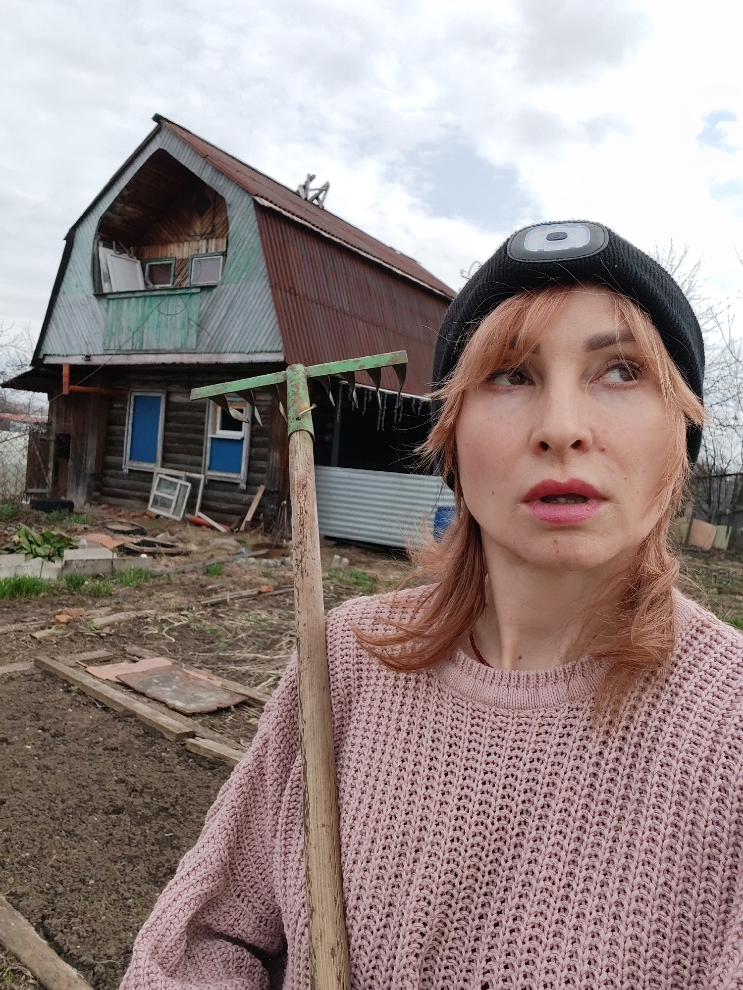The image shows a woman standing in a yard, holding a rake, with a dilapidated building in the background. The woman is fair-skinned with red hair, wearing a black beanie with a light on it, and a pink knit sweater. She is looking up and to her right with a slight expression of surprise or concern. A green rake is positioned in front of her.
The building behind her appears to be an old, wooden structure with a rusty metal roof. Some windows are boarded up, and the building looks weathered and in disrepair. The yard is overgrown with grass and scattered debris.
The image was taken in the city of Chaikovsky, Russia.