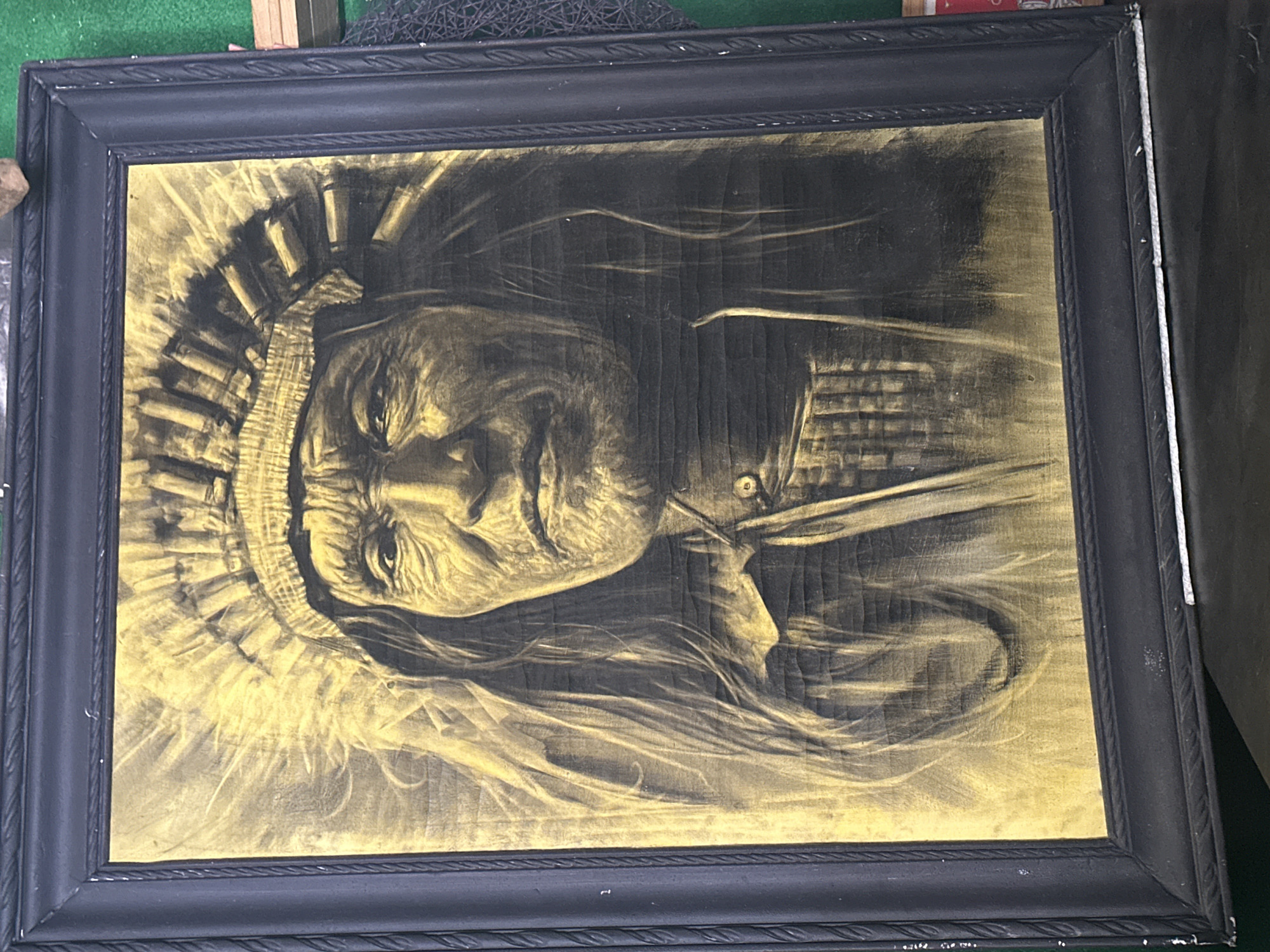 The image shows a framed artwork depicting a portrait of a Native American man in a headdress. The artwork is rendered in a gold and black palette, possibly on wood. The frame is black and ornate. The location is unknown and there is no indication of Andreyevka, Russia.