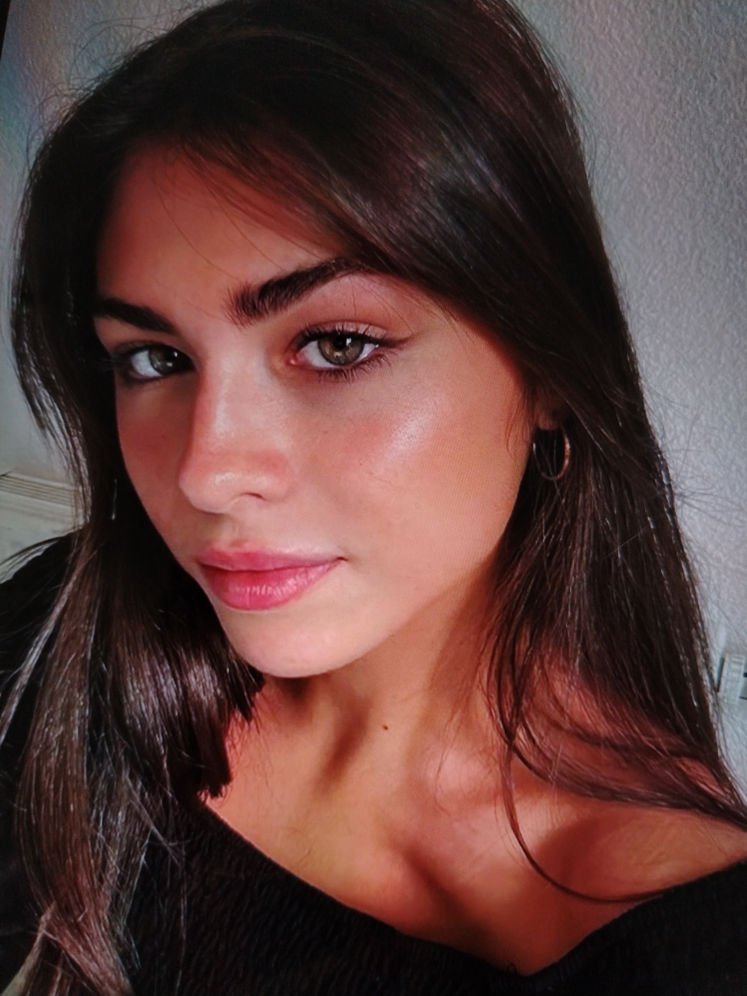 The image is a close-up, headshot of a young woman with long, dark brown hair. She has large, expressive eyes with a greenish-brown hue, defined eyebrows, and full lips. Her skin appears smooth with a subtle glow, and there is visible blush on her cheeks. She is wearing a subtle hoop earring in her right ear and a dark, off-the-shoulder top. The background is a plain, textured, light-colored wall. The lighting suggests an indoor setting, possibly a home environment. There is no indication of the city of Stavropol, Russia, or any specific location within it.