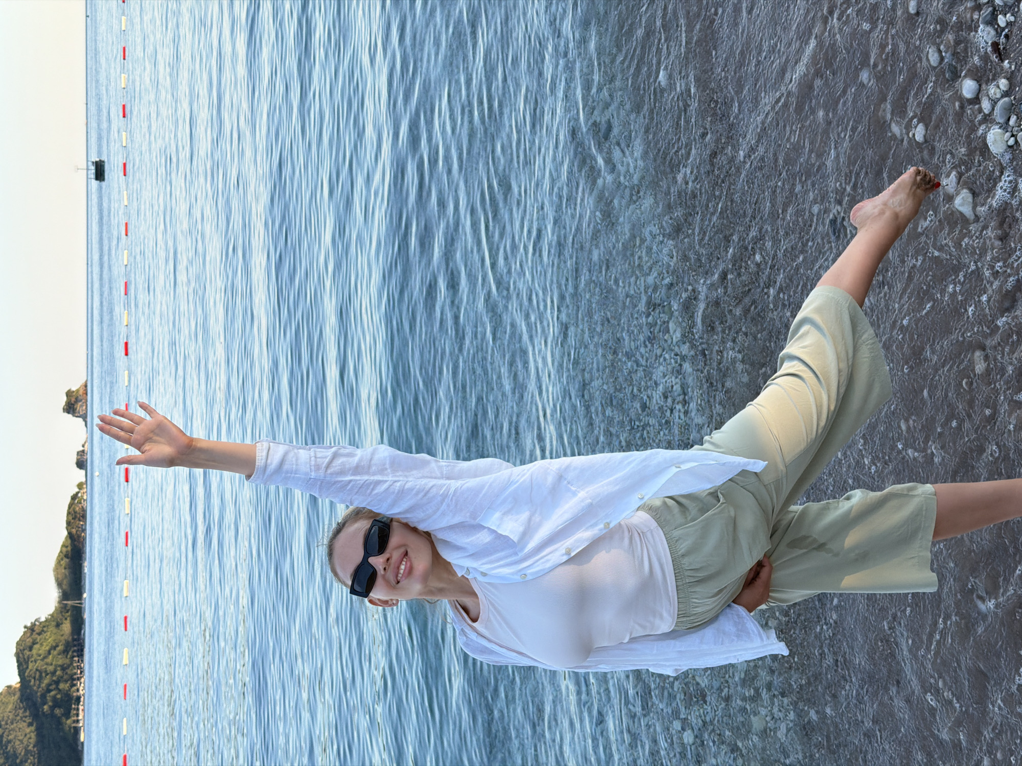 A young woman is standing on a pebble beach, with the sea in the background. She is wearing a light-colored top, a white open shirt, and light green pants. She has sunglasses on and her right arm is raised in the air, with her hand open as if waving. Her left leg is extended behind her. The water is calm with gentle ripples. In the distance, there are islands with greenery and some buildings. A rope barrier with buoys stretches across the water. The sky is clear and bright. The scene appears to be in a coastal area of Budva, Montenegro, likely a beach.