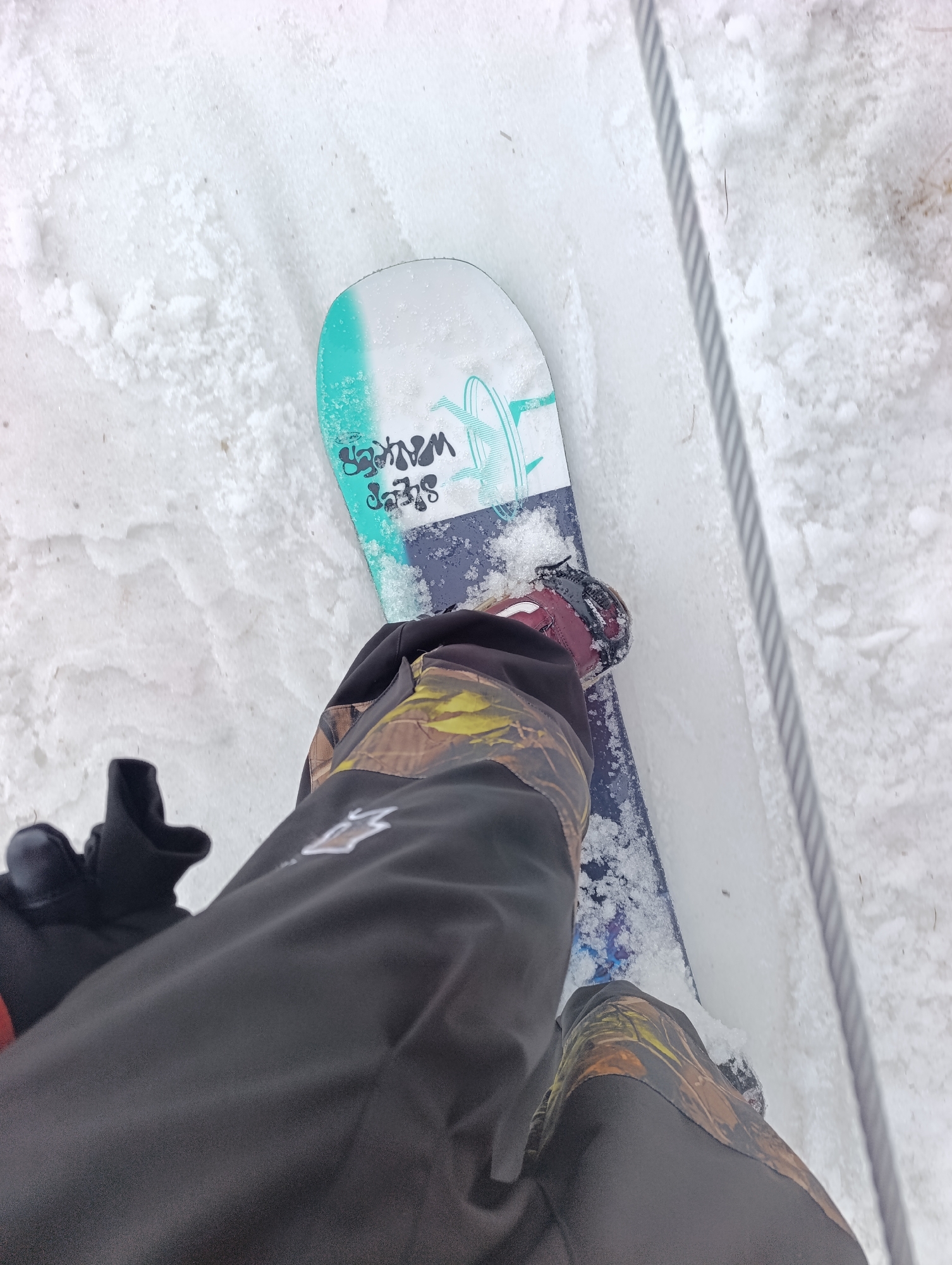 A first-person perspective photograph shows a snowboarder standing on a snowboard in the snow. The snowboard is white at the top and fades into blue toward the bottom. The snowboard has text that reads "SHEP WALKER." The snowboarder is wearing a black and camouflage snowsuit with black gloves. Snow covers the ground surrounding the snowboard. A metal cable runs parallel to the edge of the snowboard. The location is outdoors and covered in snow, potentially in a mountainous or ski resort area. There is no discernible information in the image to identify its specific location in Krasnoyarsk, Russia.