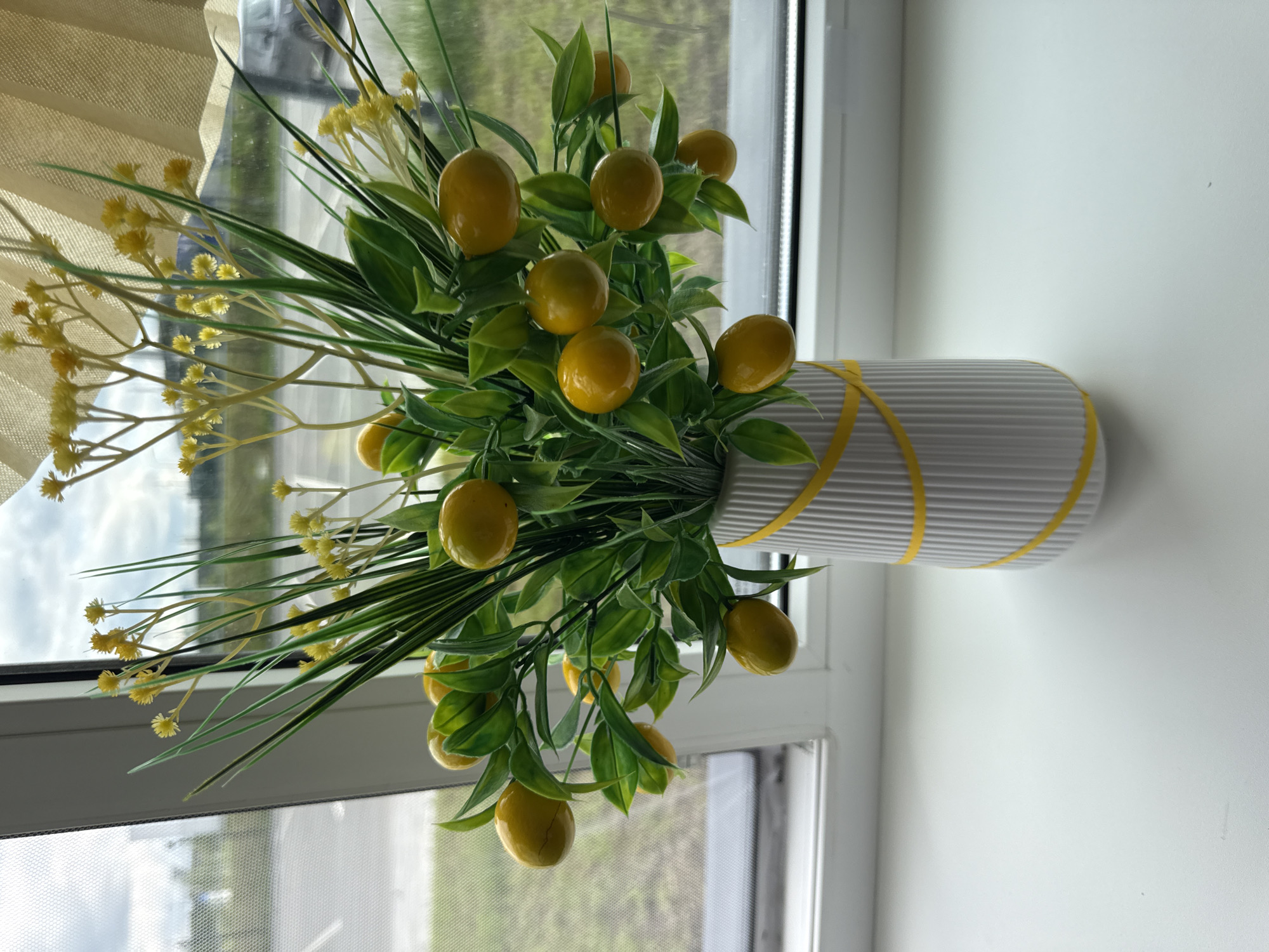 A close-up, eye-level shot shows a decorative vase filled with artificial fruit and flowers placed on a white windowsill. The vase is white with vertical ridges and is adorned with two yellow ribbon stripes. It contains a bouquet of faux lemons and small yellow flowers interspersed with green foliage and grass-like stems. The background is a window with a view of an outdoor environment, including a road and some greenery, under a cloudy sky. The image does not depict any specific location in Yekaterinburg, Russia, nor does it include any people or actions.