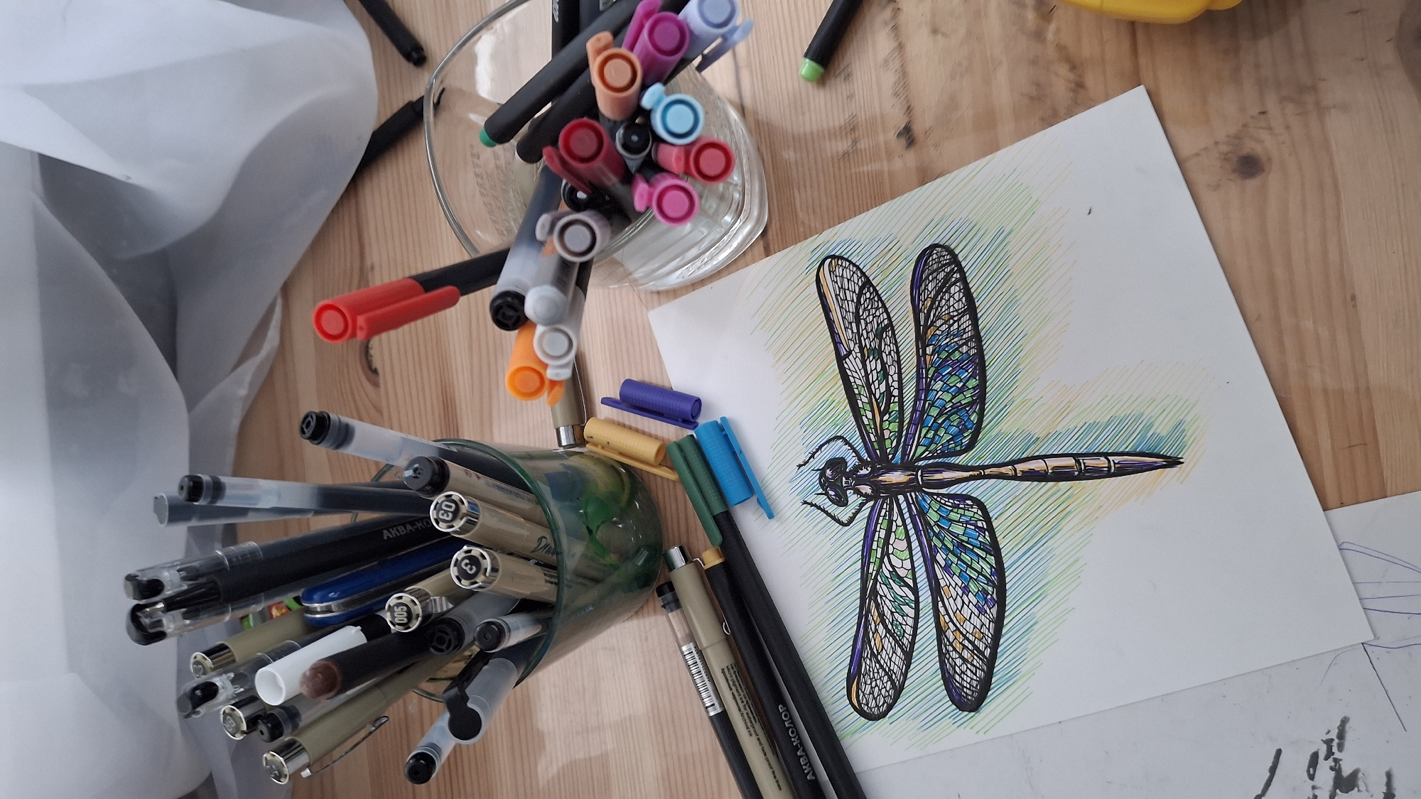 This is a top-down, eye-level shot of a wooden table, likely in a home or studio environment, located in Kempelevo, Russia. The primary focus is a drawing of a dragonfly, rendered with intricate linework and filled with colored markers. The dragonfly is positioned on a white sheet of paper, which is surrounded by an assortment of drawing tools.
Several cups and containers overflow with pens, markers, and fineliners of various colors and brands, including some with Russian labeling. Some of these writing instruments are upright in the containers, while others are scattered on the table. Marker caps are also visible, some attached and some loose. The drawing itself shows the dragonfly's wings segmented and filled with a mosaic of colors, predominantly blues and greens, with accents of yellow and purple. The body is detailed with shading and metallic-looking highlights.
The background is softly blurred, revealing a sheer, white fabric draped behind the drawing supplies, suggesting a window or curtain. The wooden surface of the table exhibits a natural grain and texture. The lighting appears to be natural, possibly from an overhead source, casting subtle shadows on the table and objects. There are no people or animals depicted in the scene. The overall impression is that of an artist's workspace, actively being used for a creative project.