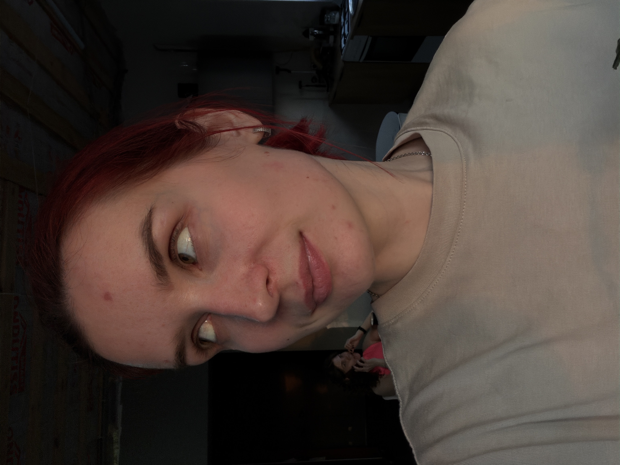 The image displays a young woman with red hair and fair skin, captured in a close-up, rotated, selfie-style photograph. She is wearing a light-colored (possibly beige or light grey) crew-neck t-shirt and a thin silver chain necklace. A small, sparkly earring adorns her left ear. Her eyes are turned upwards and to her right (viewer's left), with a slight hint of white visible beneath her irises, and a subtle smirk plays on her light pink or naturally colored lips. A few small red blemishes are visible on her face and neck.
The setting is indoors, appearing to be a casually furnished room with dim lighting, suggesting evening or night. The left side of the background reveals what looks like wooden wall panels, possibly unfinished or insulation, with some faint, indiscernible red text. The right side of the background shows dimly lit furniture and what appears to be electronic equipment. A large, dark rectangular screen, likely a television, occupies the lower right, reflecting a secondary figure, possibly the same woman, wearing a bright pink top.
The woman's expression conveys a candid, possibly playful or amused mood. The overall scene suggests a personal moment in a casual residential environment in Chaikovsky, Russia.