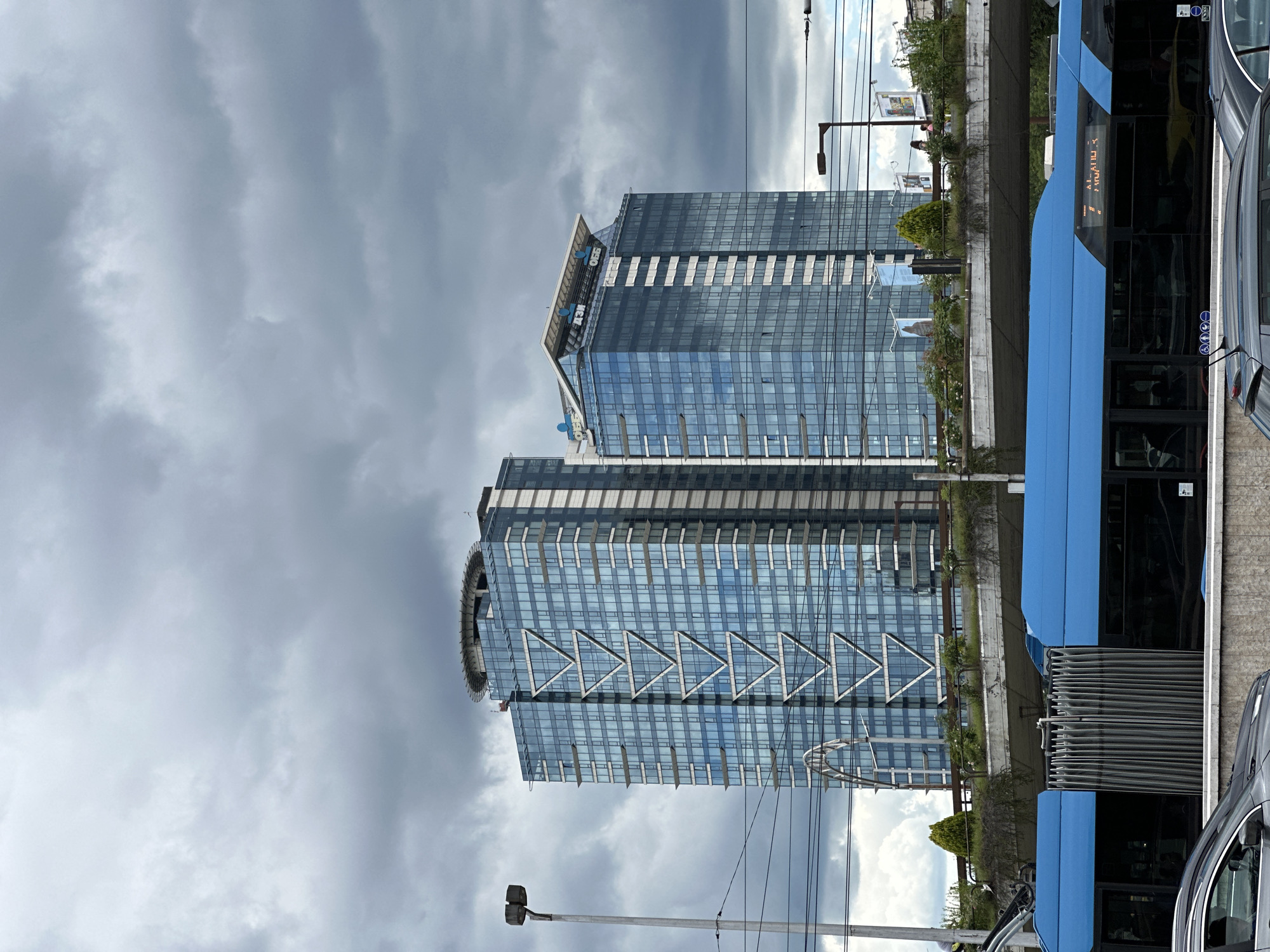 The image captures a view of the Capital Fort building complex in Sofia, Bulgaria, under a cloudy sky. In the foreground, a bright blue tram or bus, likely a public transport vehicle, is visible. The tram features a segmented body with dark windows and a distinctive accordion-style connector between sections. A portion of a dark-colored car is also present in the lower left corner.
Behind the tram, an elevated green space with trees and vegetation lines a concrete overpass. Further in the background, two modern glass-facade towers, characteristic of the Capital Fort complex, dominate the skyline. The left tower displays a prominent V-shaped pattern in white on its blue-tinted windows. The right tower has "AZI" and "OBB" signage on its upper section, also appearing in blue. A helipad is visible on top of the left tower. Overhead, a network of electrical wires and a street lamp on a tall pole are present, indicating an urban setting. The overall scene suggests a busy urban environment on a day with overcast weather.