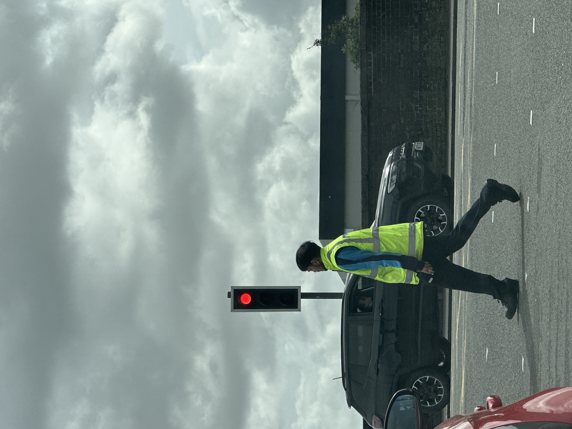 A man wearing a yellow high-visibility vest and dark clothing crosses a street in Tunbridge Wells, United Kingdom. He walks in front of two vehicles: a black SUV and a red car. A traffic light shows a red signal. The sky is cloudy. A brick wall borders the roadway.