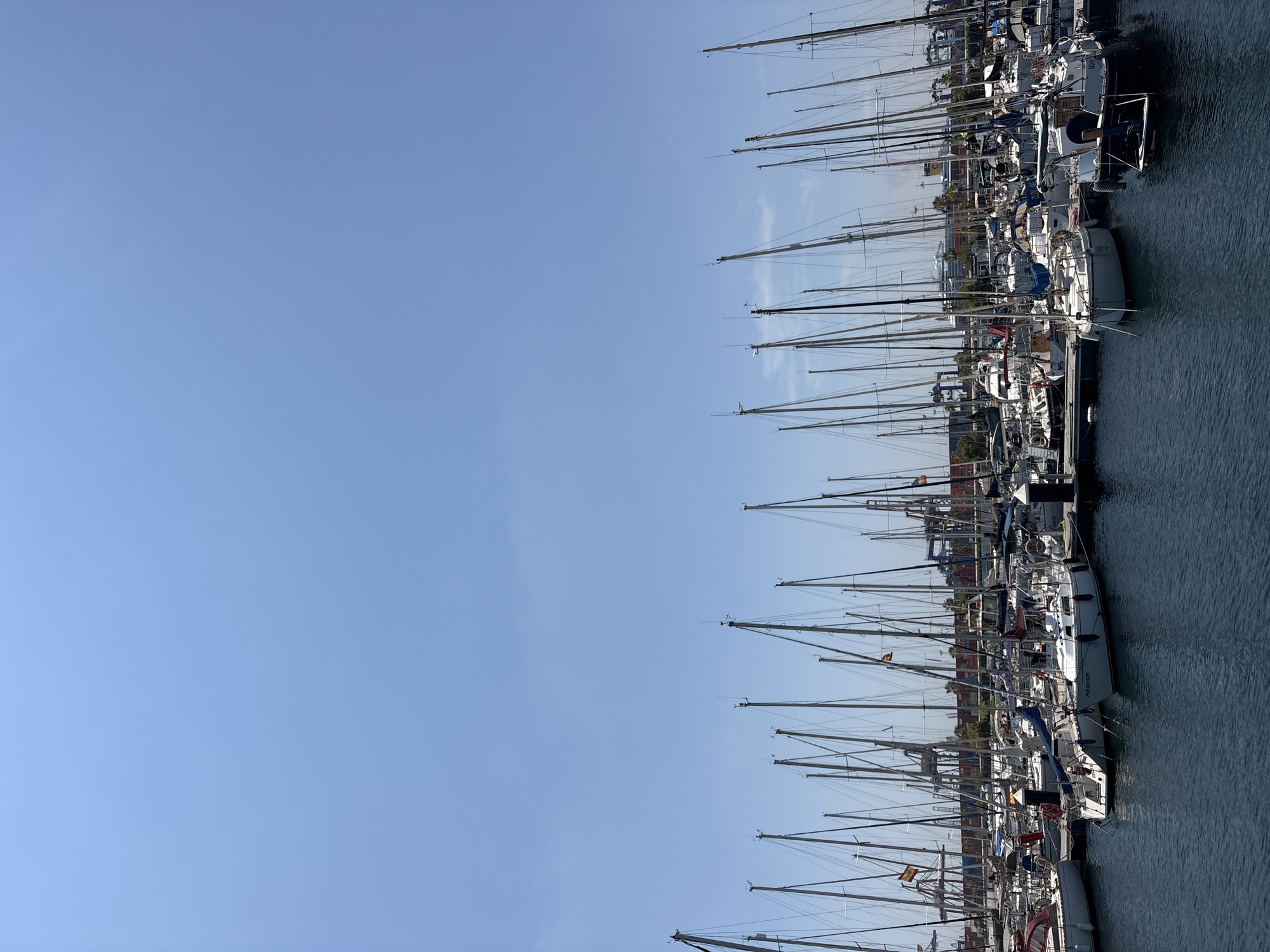 This media file depicts a marina filled with numerous sailboats docked side-by-side under a clear blue sky. The scene is likely set in Valencia, Spain, given the context. The sailboats are arranged in parallel rows, with their tall masts creating a striking visual pattern against the expansive sky. Most of the boats appear to be white or light-colored, with some sporting blue accents or sails.
The image captures a sense of quiet order, with the boats at rest in the harbor. The water is calm, reflecting the clear sky. The time of day appears to be daytime, possibly late morning or early afternoon, as indicated by the bright sunlight and the quality of the shadows. There are no people visible in the frame, emphasizing the stillness of the scene.
Some notable details include the flags displayed on some of the sailboats, suggesting different nationalities or club affiliations. One boat has the name "POEMAFLOR" visible on its hull. The masts are intricate, featuring rigging and various antennas. In the background, the outline of buildings and some greenery can be seen, providing a hint of the urban setting surrounding the marina. The overall mood is serene and peaceful, characteristic of a pleasant day at the harbor.