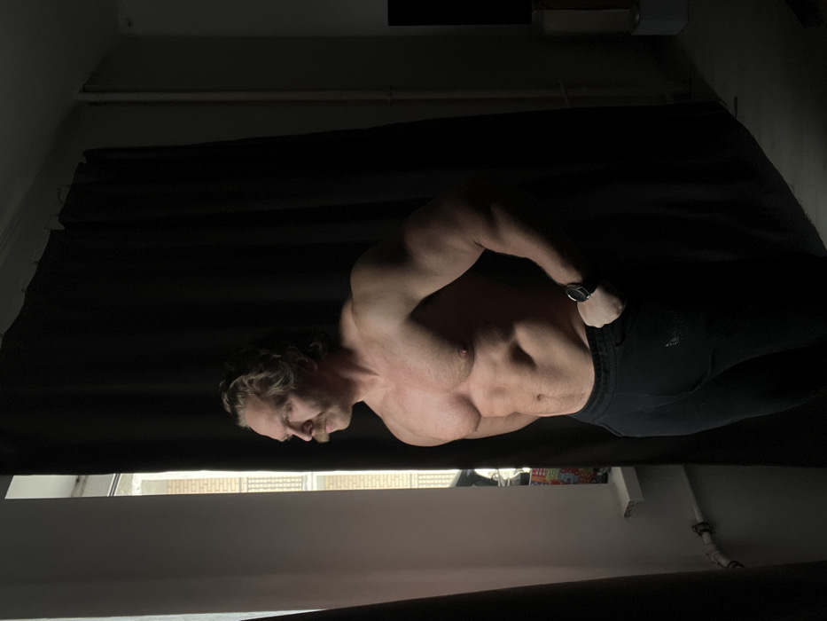 A muscular adult male is depicted standing in profile, facing left, looking out a window. He possesses fair skin, wavy light brown hair, and a prominent mustache. The man is shirtless, showcasing a well-developed physique with visible pectorals, deltoids, and abdominal muscles. He wears dark-colored sweatpants, and a dark smartwatch is present on his left wrist. His left hand is positioned near his hip. Bright natural light streams from the window on the left, illuminating his left side and creating stark contrast with the right side of his body and the interior of the room, which are in deep shadow. A black curtain is drawn across the right portion of the window and extends into the room. Several indistinct, colorful objects are visible on the white windowsill. In the dimly lit background of the room, a white pipe is visible near the ceiling, and on the far right, a dark screen and a light-colored piece of furniture are vaguely discernible. Outside the window, sections of a light-colored urban building with multiple windows are visible, consistent with residential architecture found in a city such as Saint Petersburg, Russia.