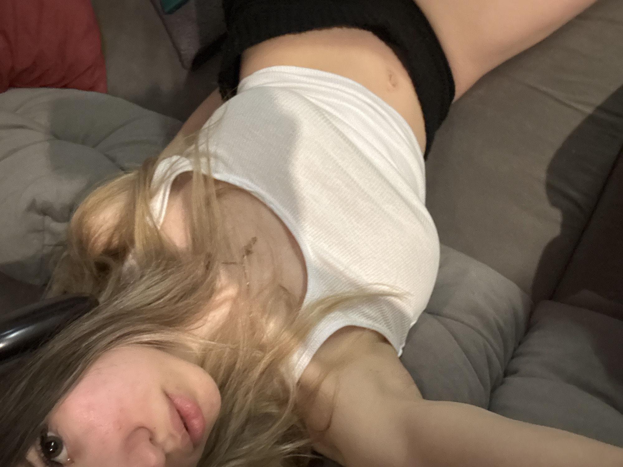 A young woman with long, light-colored hair is lying diagonally across a grey sofa, captured from a slightly elevated perspective. She is looking upwards towards the camera, with her head tilted to her left. Her hair is spread around her head and shoulders. She wears a white ribbed crop top that reveals her midriff and navel, and black shorts or underwear. Her facial expression is neutral, with her eyes open and lips slightly parted.
In the background, a large grey cushioned sofa is visible, along with a pink pillow on the left side of the frame. The lighting appears to be indoor, creating subtle shadows. The overall scene suggests a relaxed or posed moment, likely a selfie taken indoors. No visible text is present in the image.