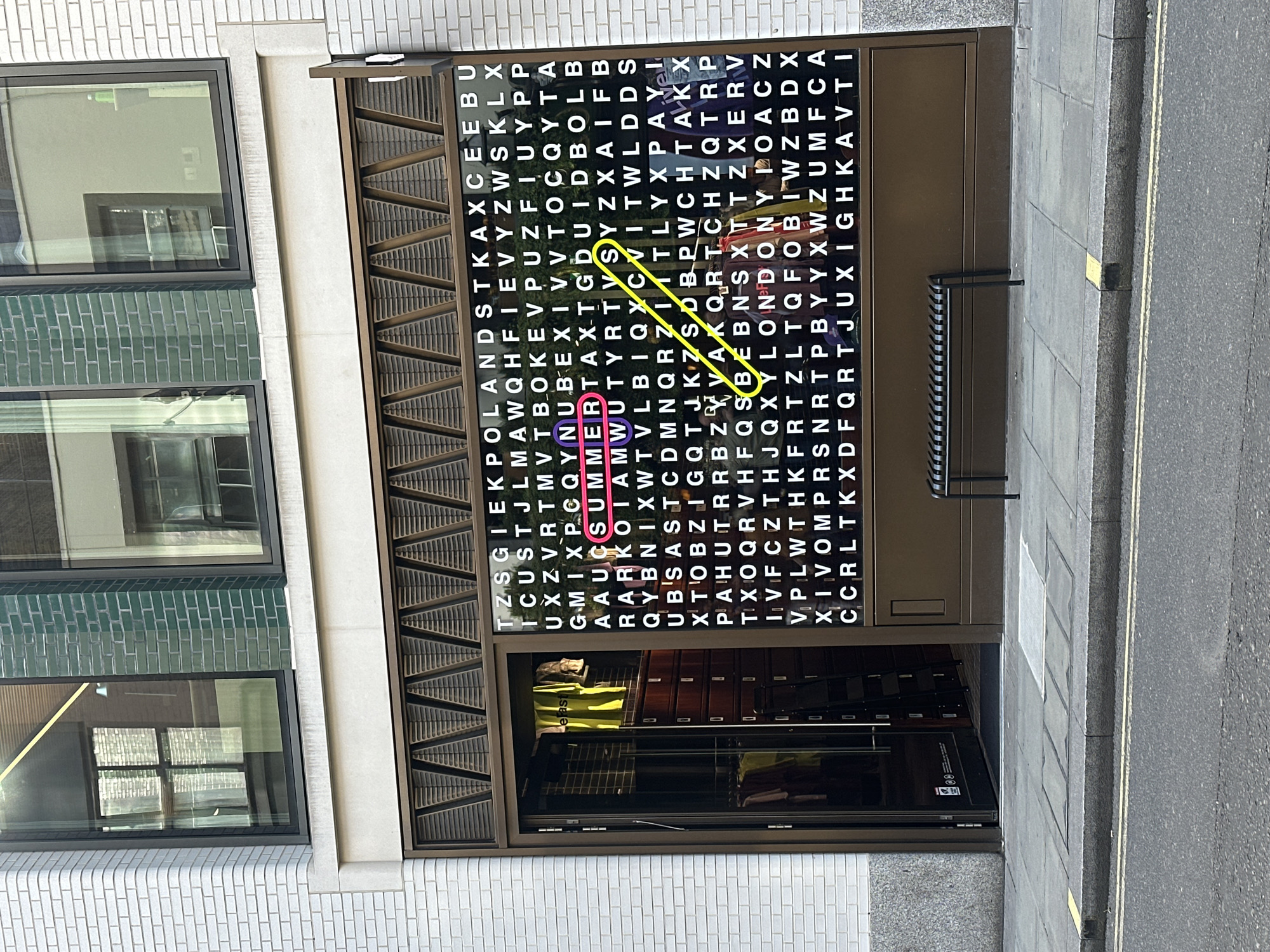 The image captures the front of a retail establishment, possibly a clothing store, in London, United Kingdom. The store's entrance features an unusual display. The display is constructed like a word search puzzle with letters arranged in a grid, highlighting the word "SUMMER" with a yellow line drawing over the letters. A large pink paperclip is clipped onto the word "SUMMER". A display can be seen in the shop window, showcasing clothing items and cabinets with drawers. Outside the store, a grey sidewalk is visible. The store is constructed with light colored brick or tile, and has multiple windows.