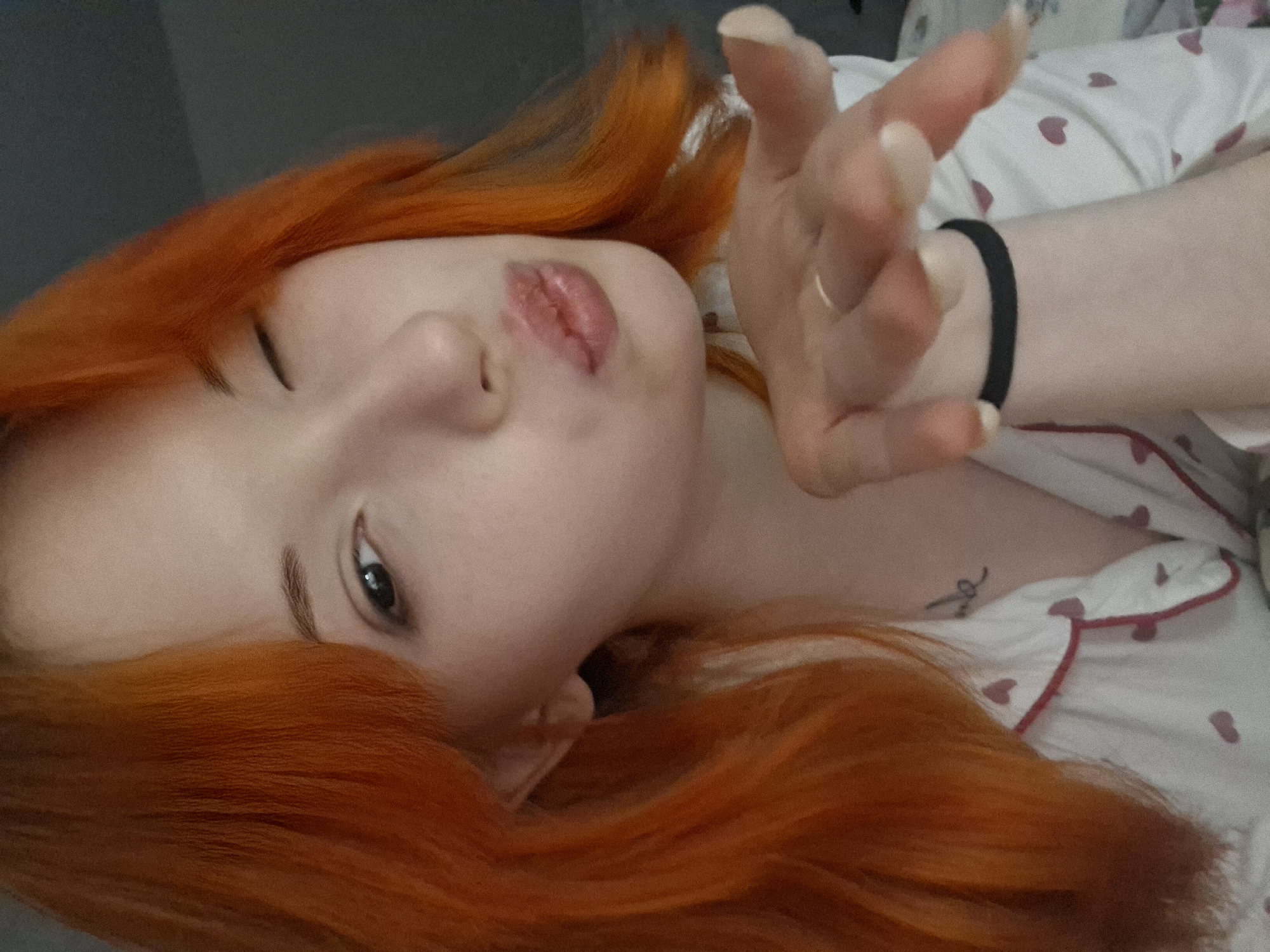 An image captures a person, likely female, with prominent, long, wavy, bright orange-red hair, posing in an indoor setting. The individual's face is shown at a tilted angle, with one eye partially open and looking directly at the camera. Their lips are pursed in a "kiss" gesture, conveying a playful and affectionate mood.
The person has light skin and distinct, dark eyebrows. Their right hand is extended towards the camera, with fingers slightly splayed. A black hair tie is visible on their right wrist, and a simple ring is worn on one of their fingers. Their fingernails are long and light-colored. They are wearing a white top or pajamas adorned with a pattern of small pink or red hearts and featuring red piping, suggesting a comfortable, personal environment. A small, indistinct tattoo is visible on their upper chest or shoulder area, though its specific design or any text is too blurry to discern.
The background is soft and slightly out of focus, consisting of a dark, muted area on one side and the heart-patterned fabric on the other, reinforcing a cozy, intimate atmosphere. The scene suggests a relaxed moment, possibly in a bedroom in Chaikovsky, Russia.