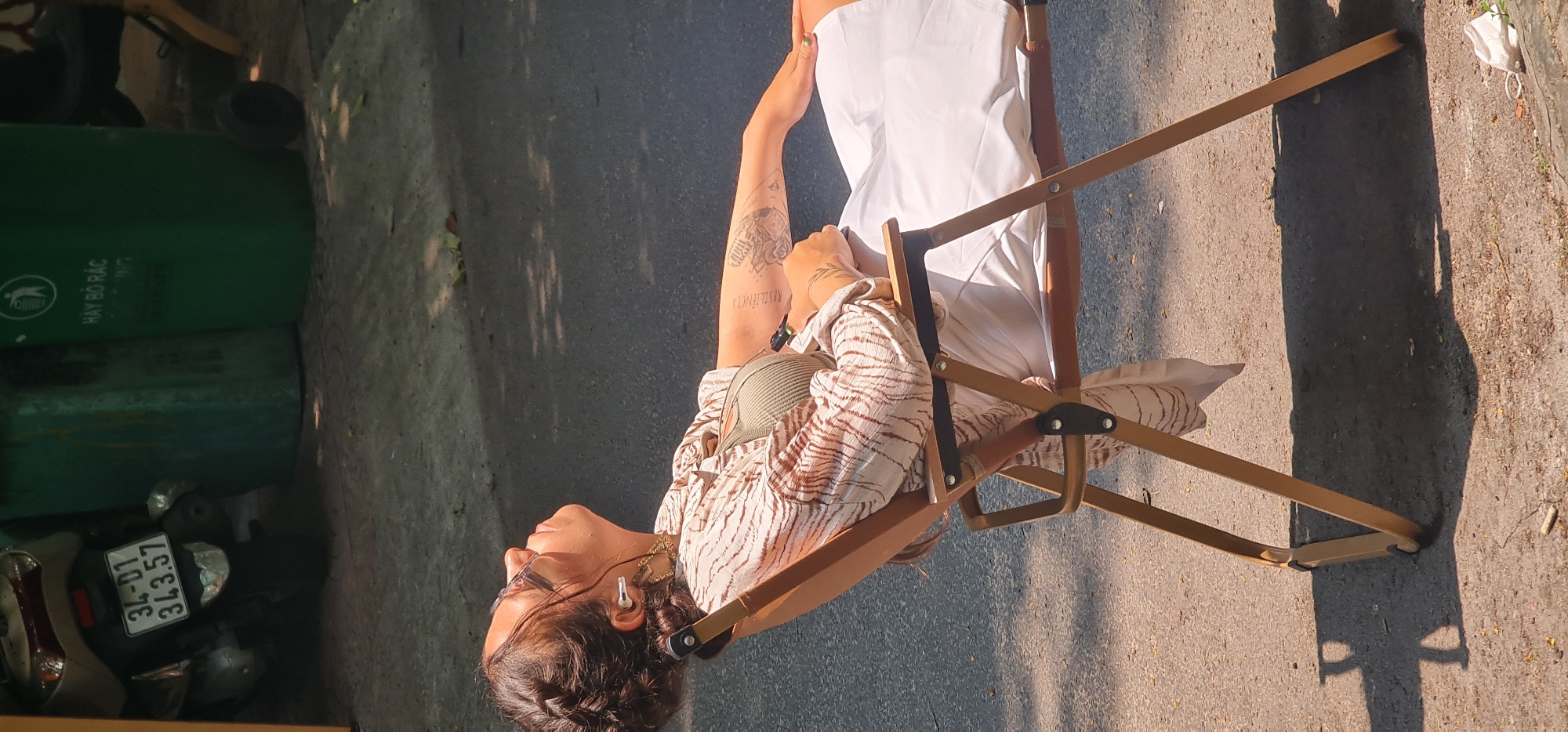 A medium shot shows a person sitting in a wooden chair on an asphalt street in an urban setting. The person has dark hair braided back, wears glasses, and has several ear piercings. The person wears a light brown and white patterned shirt and a light green top. The person's left arm has a tattoo on the forearm and another on the upper arm. The person sits against the back of the chair, with their head tilted back. They wear a white garment over their legs. The chair is a folding wooden chair. In the background, there is a large green trash bin and a parked scooter with a license plate that reads "34-01 343.57". The location is possibly in the city of Đà Nẵng, Vietnam.