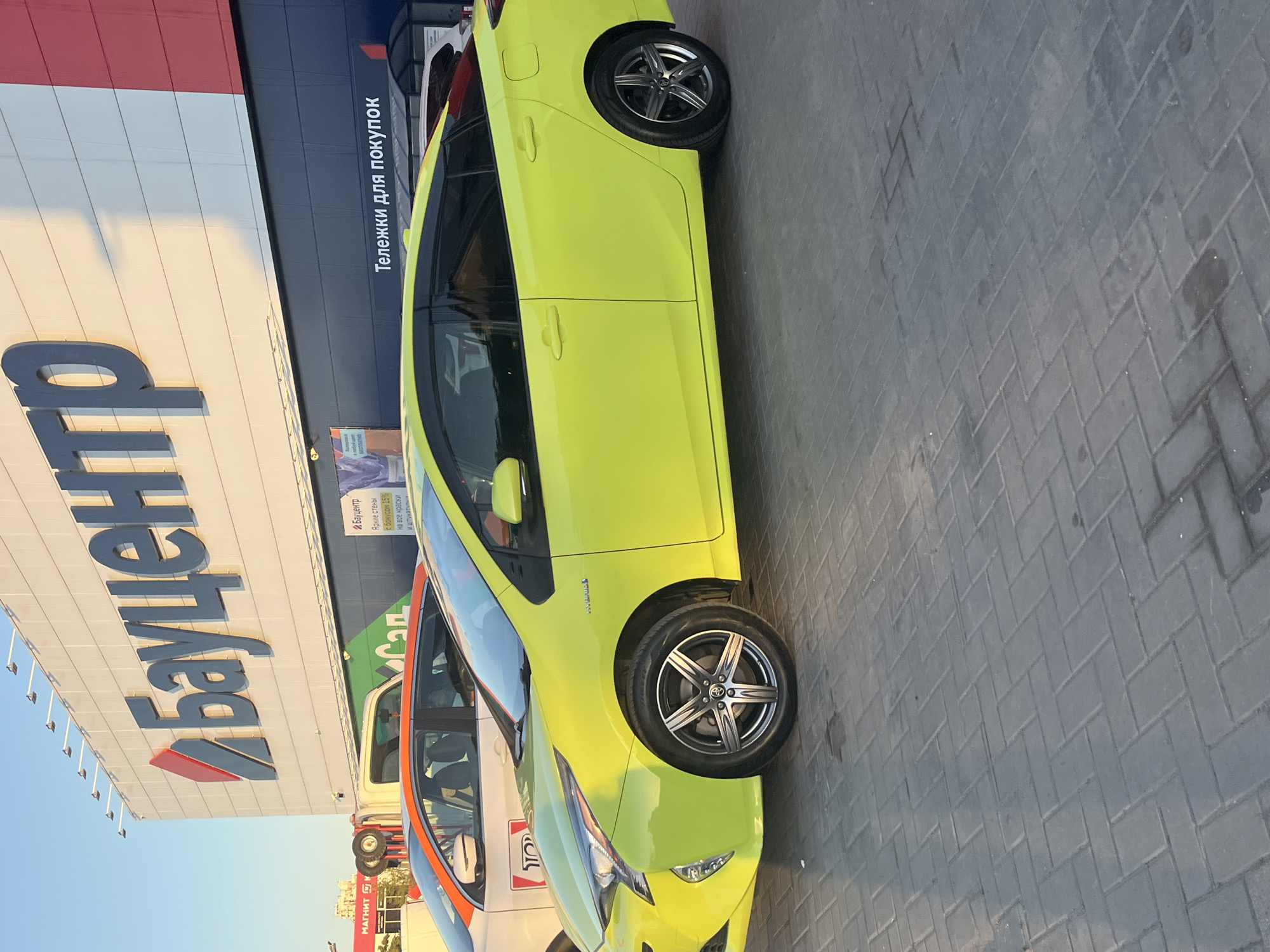 The image shows a bright yellow hybrid car parked on a paved area in front of a building with large blue and red signage. The building appears to be a retail store, with the name "Бауцентр" (Baucenter) prominently displayed in large blue letters. To the left of the yellow car, another car, primarily white with an orange stripe, is also visible. Above the cars and to the left, the text "Тележки для покупок" (Shopping carts) is visible in white letters on a dark background.
The lighting suggests it is daytime, with shadows cast by the cars and the building. The sky is a clear blue, indicating fair weather. The ground is paved with rectangular bricks laid in a herringbone pattern. The overall scene conveys a sense of everyday commercial activity.
Visible text includes "МАГНИТК", "Бауцентр", "Яркие стены с бонусом 15% на все краски И штукатурни в любой цвет БЕСПЛАТНО", and "Тележки для покупок". These texts suggest a shopping environment, possibly a hypermarket or hardware store. The presence of shopping carts indicates that customers are likely shopping or have just finished shopping. The "Baucenter" branding is consistent with a home improvement or construction materials store.