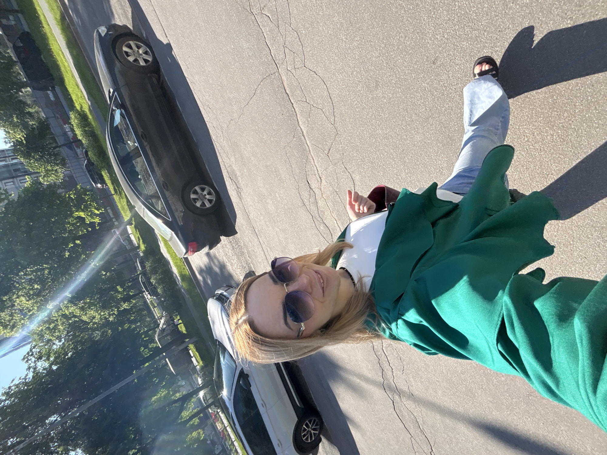 The image captures a bright, sunny outdoor scene, featuring a young woman posing for a selfie or photograph on the side of a paved road. The woman, with light skin and blonde hair, is smiling and wearing large, dark sunglasses. She is dressed in a white top, a loose dark green jacket or cardigan, and light blue jeans, paired with black sandals. Her right hand is raised, possibly forming a thumbs-up gesture near her chest.
The setting appears to be an urban or suburban street. To her left, two cars are parked along the curb: a dark-colored sedan is prominent in the foreground, with a lighter-colored vehicle visible behind it. The road surface is asphalt, showing some cracks. Beyond the parked cars and the curb, a line of green trees and foliage extends, with structures, possibly buildings, faintly visible in the background through the trees.
The time of day is clearly daytime, characterized by bright sunlight that casts distinct, long shadows from the woman and the cars, suggesting either morning or late afternoon. A visible sun flare adds to the bright conditions. The woman's smile conveys a happy or pleasant mood. There is no visible text in the image. The specific location is not identifiable from the visual information provided.