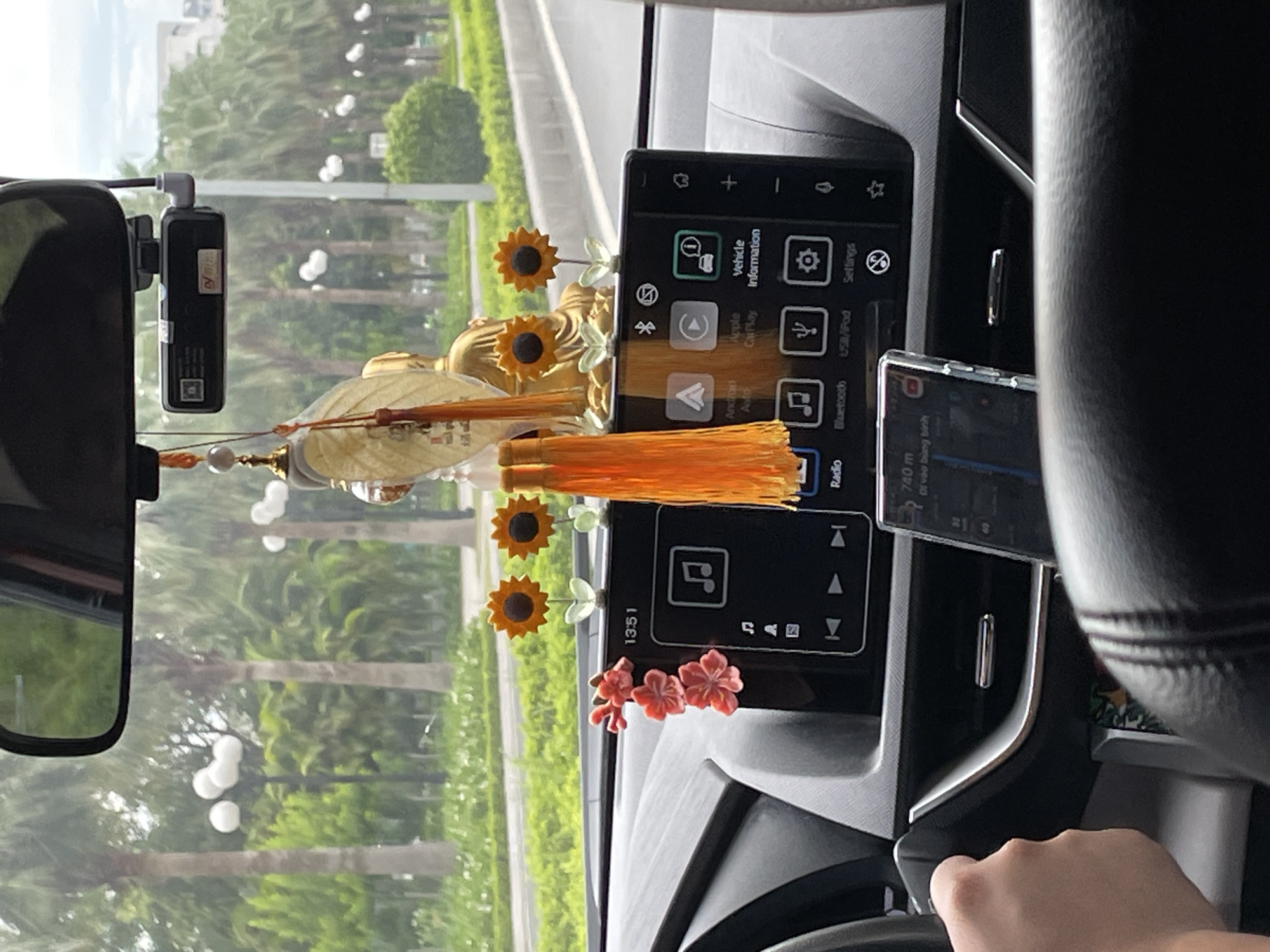 The image shows the interior of a car with a person's hand on the steering wheel. The car's dashboard is visible, featuring a central infotainment screen displaying various icons like "Radio," "Bluetooth," "USB/iPod," and "Settings." The screen also shows the time as "13:51." There are decorative items on the dashboard, including small sunflower ornaments and a cluster of pink flowers. A pendant with a golden Buddha-like figure and orange tassels hangs from the rearview mirror. A dashcam is mounted above the rearview mirror. Outside the car, palm trees line a street, suggesting a tropical or subtropical urban environment. The presence of Vietnamese text on the phone screen, "740 m" and "Đi vào vùng bình", along with the architectural style visible in the background, is consistent with Hà Nội, Vietnam.