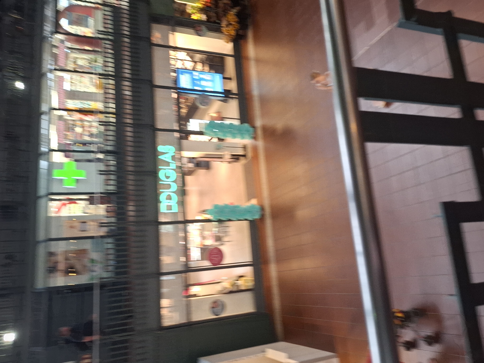 This image depicts an interior view of a shopping mall. In the upper section of the image, a store with a bright green plus sign is visible, suggesting it could be a pharmacy or a health and beauty store. Below this, a store with the prominent, illuminated "DOUGLAS" logo is seen. Douglas is a well-known international retailer of beauty products. The mall appears to have multiple levels, with a railing in the foreground suggesting the viewpoint is from an upper floor. The flooring is wooden. In the background, several people are visible, though their features are indistinct due to the image's blurriness. One person, a child, is standing on the wooden floor in the lower right portion of the image. The overall lighting suggests an indoor environment, possibly during evening hours. The specific location in Hamburg, Germany, cannot be determined from the visual information alone.