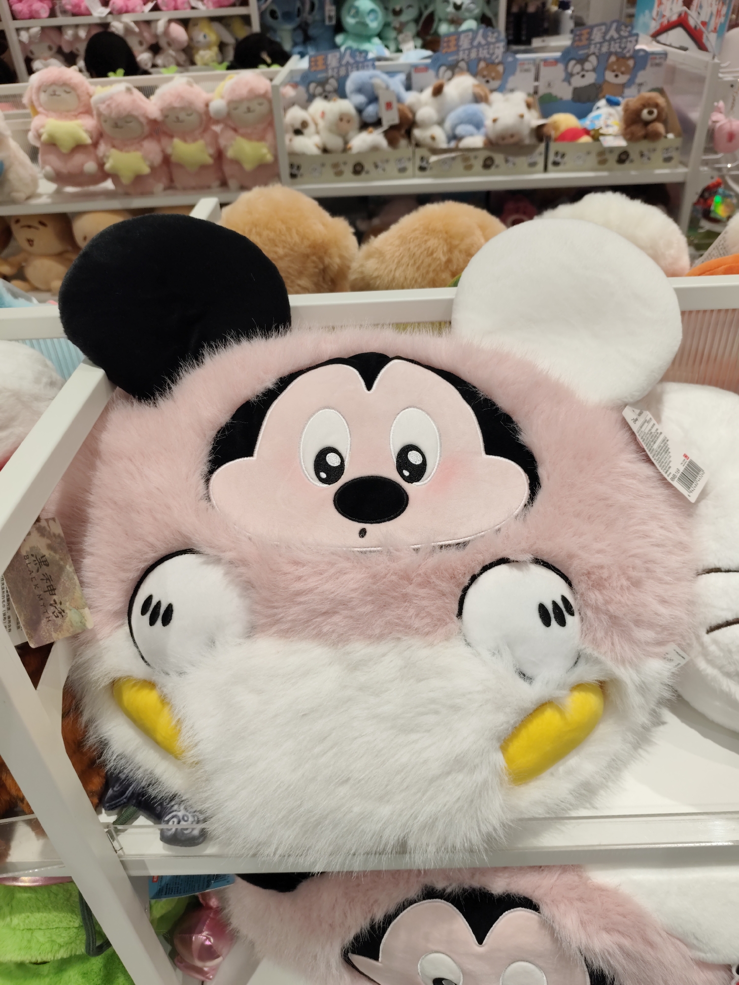 A fluffy, pink Mickey Mouse plush toy is displayed on a white shelf in a store. The plush toy has large, round black ears, a pink face with large white eyes and black pupils, and rosy cheeks. Its body is white and fluffy, with yellow accents representing its paws or shoes. In the background, other stuffed animals and merchandise are visible on shelves, suggesting a retail environment. The overall impression is that of a well-lit store selling children's toys. The scene is located in Daqing, China.