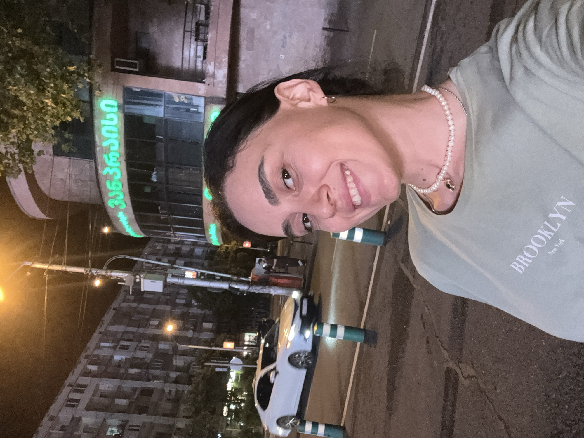A young woman is taking a selfie at night on a street in Tbilisi, Georgia. She is wearing a light green t-shirt with "BROOKLYN New York" printed on it and a pearl necklace. Behind her, a white car is driving on the road, and there are bollards with green and white stripes lining the street. In the background, a building with green neon signage in Georgian script is visible, along with a multi-story apartment building and a lamppost. The scene suggests a casual evening in the city.