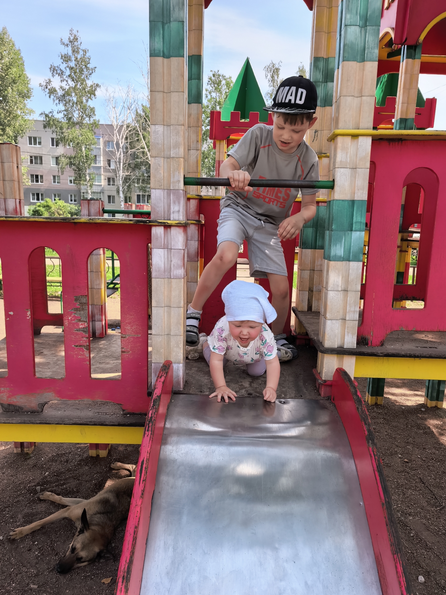 A boy in a gray t-shirt and shorts with the word "MAD" on his black cap, and a baby wearing a light blue headscarf and a floral print onesie, are on a colorful playground slide. The boy is standing at the top of the slide, holding a horizontal bar. The baby is at the bottom of the slide, crawling towards the boy with an excited expression. A dog is lying down on the ground to the left of the slide. The playground structure is red, yellow, and green, with square beige and green pillars. In the background, there is a multi-story apartment building and trees. The scene appears to be in the city of Sterlitamak, Russia, based on the surrounding architecture and vegetation. The ground is covered with dirt and small stones.