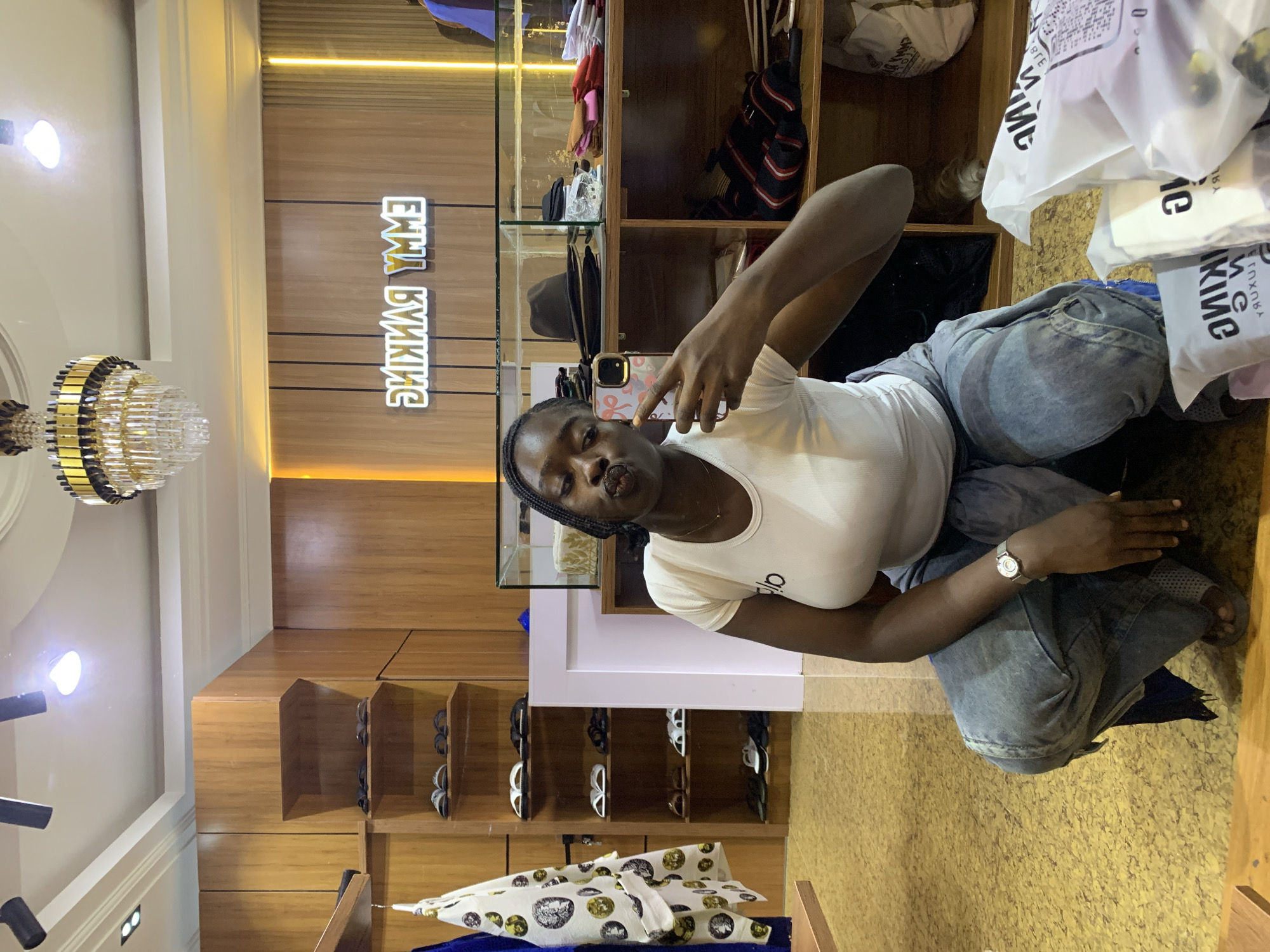 A young Black woman is taking a selfie in a retail store. She is wearing a white t-shirt, baggy denim jeans, and a wristwatch. The store appears to be a clothing boutique, with wooden shelves displaying shoes, hats, and folded clothing. The woman is squatting down, with her phone held up to take the photo. The background shows a bright, modern interior with a chandelier on the ceiling and a neon sign that reads "EMMY RANKING". The store appears to be well-lit and organized. The woman's facial expression is playful, with her lips pursed as if blowing a kiss. She is the sole subject of the image and is the focus of the selfie.