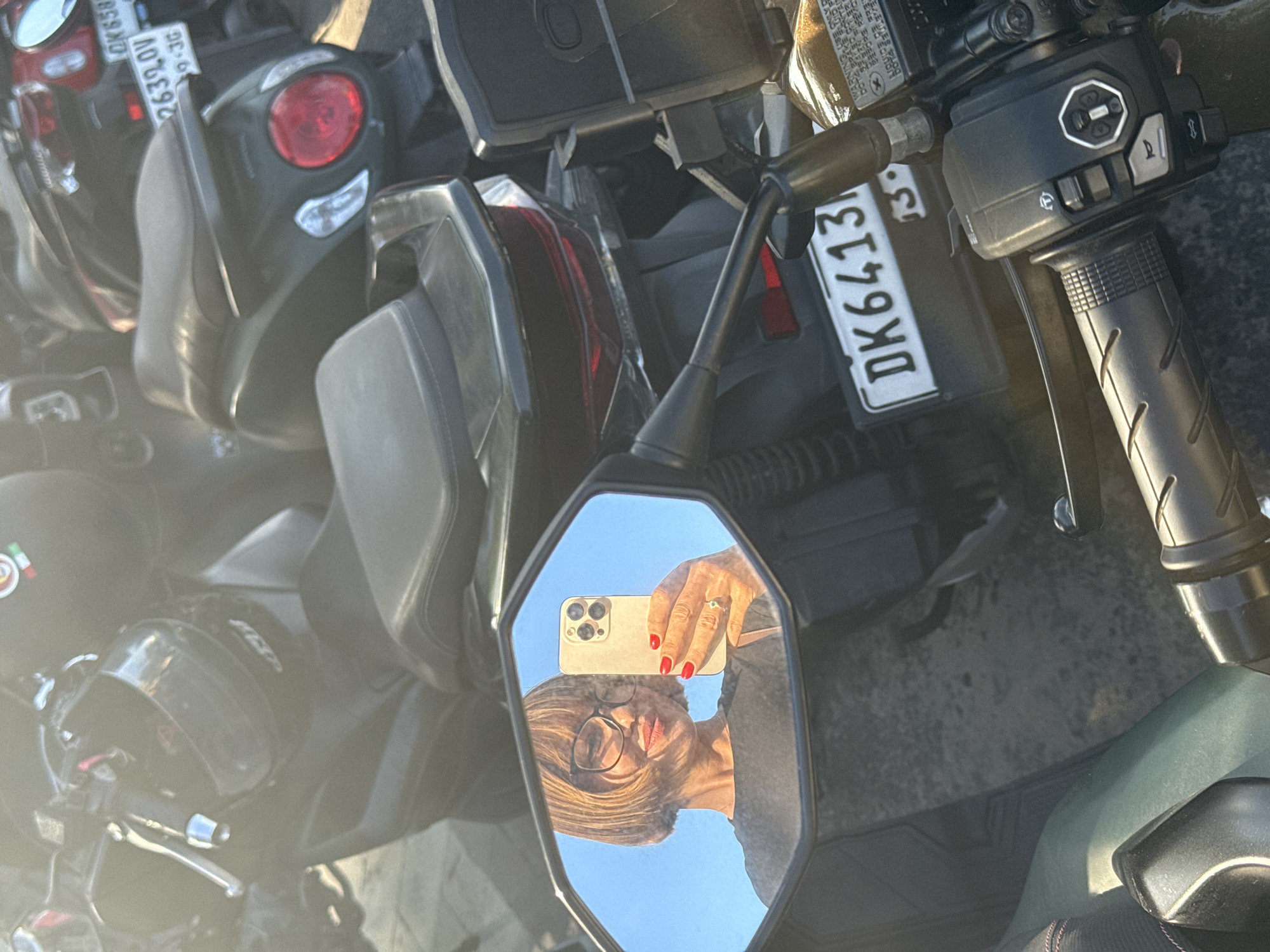The image shows a scene in Kuta, Indonesia. It is a close-up shot taken from the perspective of a motorcycle rider, focusing on the motorcycle's side mirror. The mirror reflects a woman holding a smartphone, seemingly taking a selfie. Her reflection also reveals that she is wearing glasses and has short blonde hair and red lipstick. Her hand is visible in the reflection, showing red nail polish and a ring.
The background of the image consists of other motorcycles parked closely together. Visible details of the motorcycles include taillights, seats, handlebars, and license plates. One visible license plate shows the designation "DK6413I3." The environment suggests a crowded parking area, likely in an urban setting. The weather is clear and sunny.