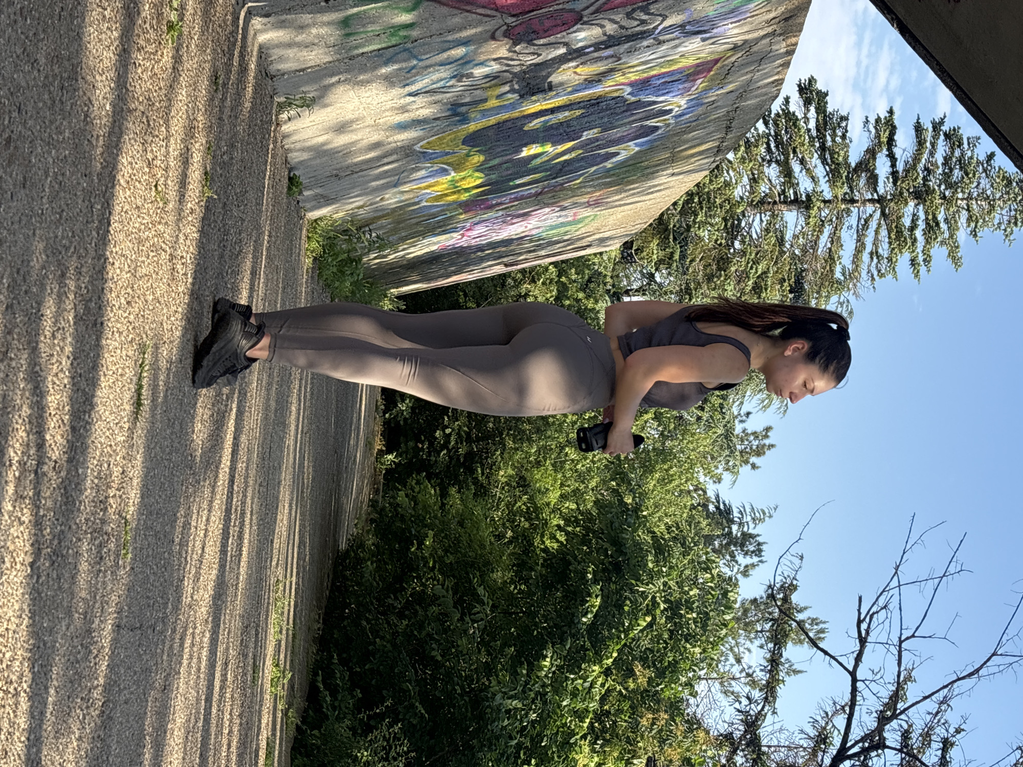 A young woman stands on a textured concrete path next to a graffiti-covered concrete wall, under a clear blue sky in an outdoor location in Varna, Bulgaria. She is wearing a dark grey athletic crop top, matching high-waisted leggings, and dark athletic shoes. Her dark hair is styled in a high ponytail. She holds a black smartphone in her right hand, positioned near her hip, and is looking downwards.
The concrete wall behind her is tall and features colorful, abstract graffiti designs. To her right, a lush expanse of green trees and bushes fills the mid-ground, extending towards the clear blue sky with scattered white clouds. The ground she stands on is light-colored and granular, with pronounced, elongated shadows stretching across it, indicating direct sunlight and possibly a morning or late afternoon setting. No readable text is visible on her clothing or the phone, though the graffiti consists of abstract patterns. The overall scene suggests an active urban-nature blend, with the woman appearing focused or contemplative.