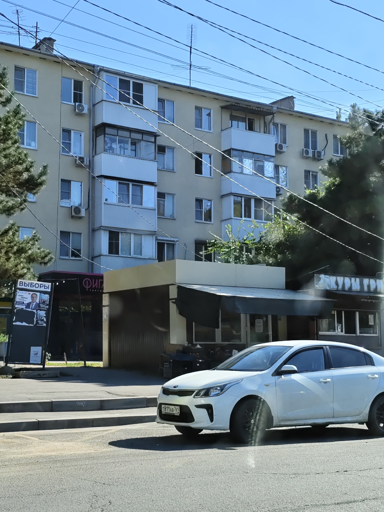 The image shows a white sedan driving on a street in Rostov-on-Don, Russia. The car has a license plate that reads "T811AB 761". In the background, there is a multi-story building with several windows and balconies. On the ground floor of the building, there are several shops, including one with a sign that says "ВЫБОРЫ" (Elections) and another with a sign that appears to read "КУРЫ ГРИЛ" (Rotisserie Chicken). There are also several power lines visible in the sky. The weather appears to be sunny and clear.