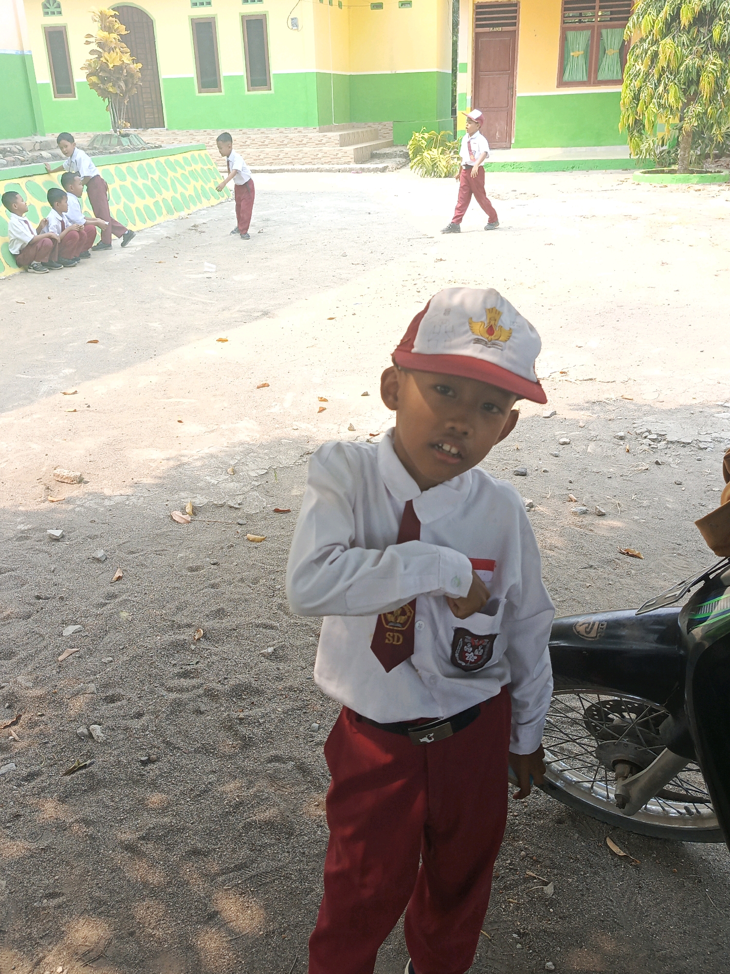 A young boy in a white school uniform with a red tie and red pants, and a red and white baseball cap stands in the foreground. He is looking at the camera with his mouth slightly open. Behind him, several other children in similar uniforms are visible in the background, some sitting on a low wall and others standing or walking. The environment appears to be an outdoor schoolyard with a sandy or gravel ground. In the far background, there is a building with yellow and green walls. The scene suggests a school day activity in Karya Dadi, Indonesia.