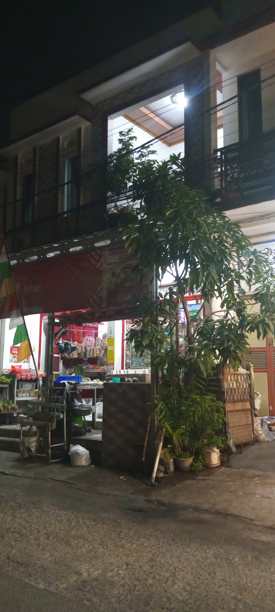 The image shows a street-level view of a small shop or convenience store at night in Setu, Indonesia. A tree with dense foliage is prominent in the foreground, obscuring a portion of the building's facade. The building appears to be two stories, with an open-air upper level visible. The shop itself has a red awning and its interior is illuminated, revealing shelves stocked with goods and a counter area. Several flags, including a green and white flag, are visible on the left side of the storefront. Various items, such as plastic bags, buckets, and what appear to be plants in pots, are scattered around the entrance and the adjacent street. The street surface is wet and reflective, indicating recent rain or cleaning. Electrical wires are strung across the upper portion of the image. The scene is dimly lit by artificial lights, characteristic of a nighttime urban environment.