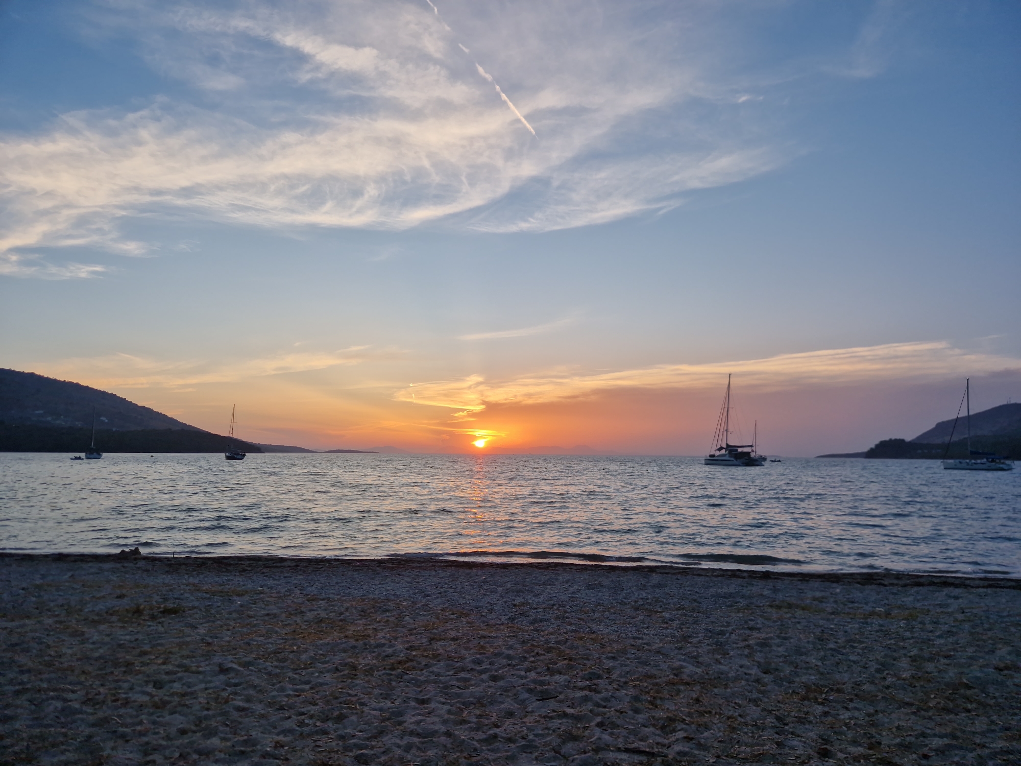 A panoramic, eye-level, long shot captures a sunset scene over the Ionian Sea from the beach in Plataria, Greece. The sky is painted with streaks of white and wispy clouds against a gradient of blue, orange, and pink hues, indicative of twilight. The sun, a bright orange orb, is positioned near the horizon, casting a shimmering reflection on the calm water. Several sailboats are visible on the sea; one large catamaran is in the middle ground, while smaller sailboats are scattered throughout. The coastline is lined with a thin strip of sandy beach in the foreground, with a gentle slope leading to the water's edge. Hills, covered in vegetation, form the distant background on the left side of the image. The overall atmosphere is serene and tranquil, characteristic of a peaceful evening by the sea.