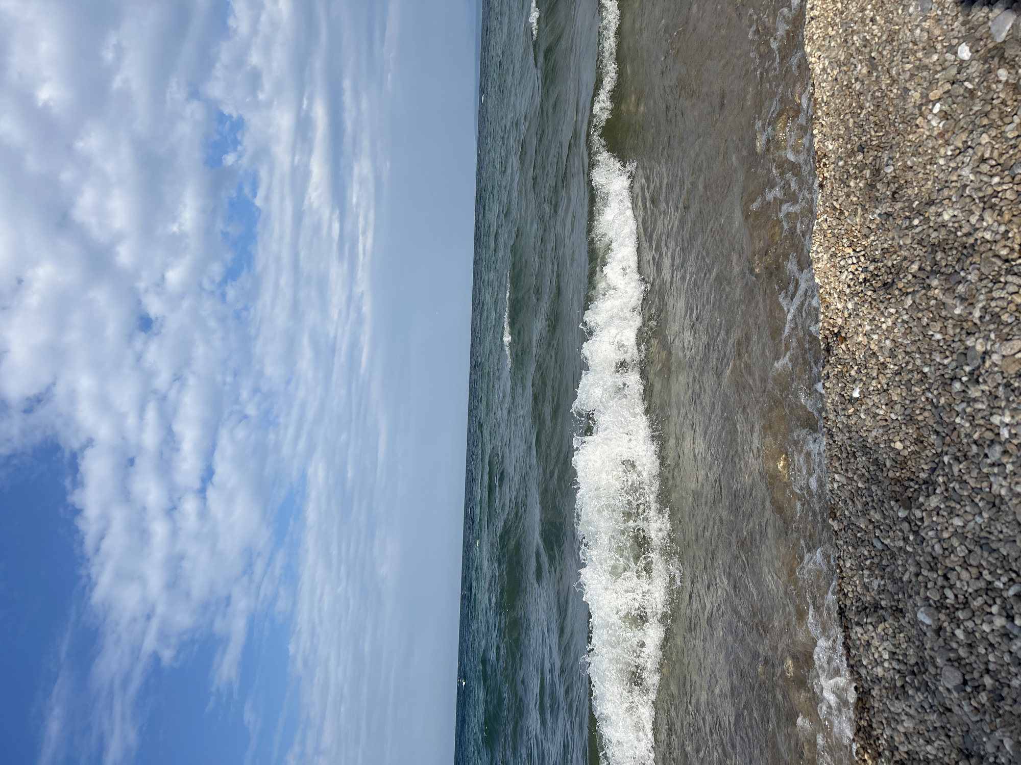 The image depicts a natural scene at the shore of a body of water. In the foreground, a pebbled beach is visible, with small, rounded stones in shades of gray and beige. The water's edge meets the pebbles, with small waves lapping at the shore, creating white foam and ripples. Further out, the water appears dark green and choppy, with larger waves rolling in. The horizon line is visible in the distance, separating the water from the sky. The sky is a clear blue with scattered white, wispy clouds. There are no people or man-made structures present in the image. The location is identified as being in the city of Tolbazikha, Russia, implying this is a natural landscape within that area.