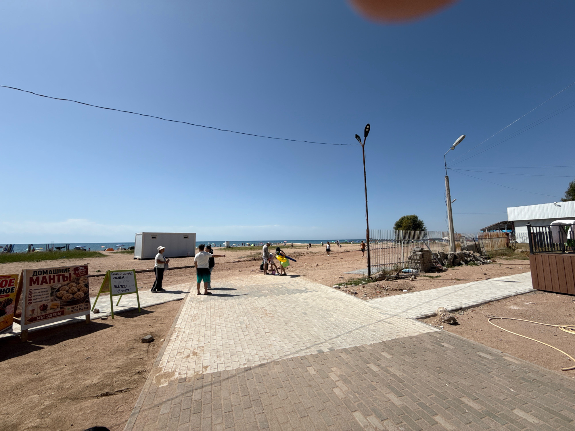 The image depicts a sunny day on a beach in Bosteri, Kyrgyzstan, with a clear blue sky overhead. The foreground features a paved walkway constructed with light-colored rectangular bricks, bordered by sandy ground. To the left, a yellow and white sign advertises "Домашние МАНТЫ" (Homemade Manty) and offers delivery services, with phone numbers listed. Further to the left, another partially visible sign features an image of fruit.
In the middle ground, a group of people is gathered on the sandy beach. Two individuals, a man in a white t-shirt and shorts and another in a white cap and dark clothing, are standing near a white portable building. Another group of people, including a child in yellow attire, are interacting near the water's edge. Beyond them, the beach extends towards the sea, with numerous beach umbrellas and sunbathers dotting the landscape. A fence separates the beach area from a rougher patch of ground with some construction debris.
In the background, the calm, blue water of a large lake stretches to the horizon. Several utility poles with wires are visible, including one with a modern street lamp. A white building with a dark roof is situated on the right side of the image, with a wooden-paneled section. The overall scene suggests a recreational area on a warm day.