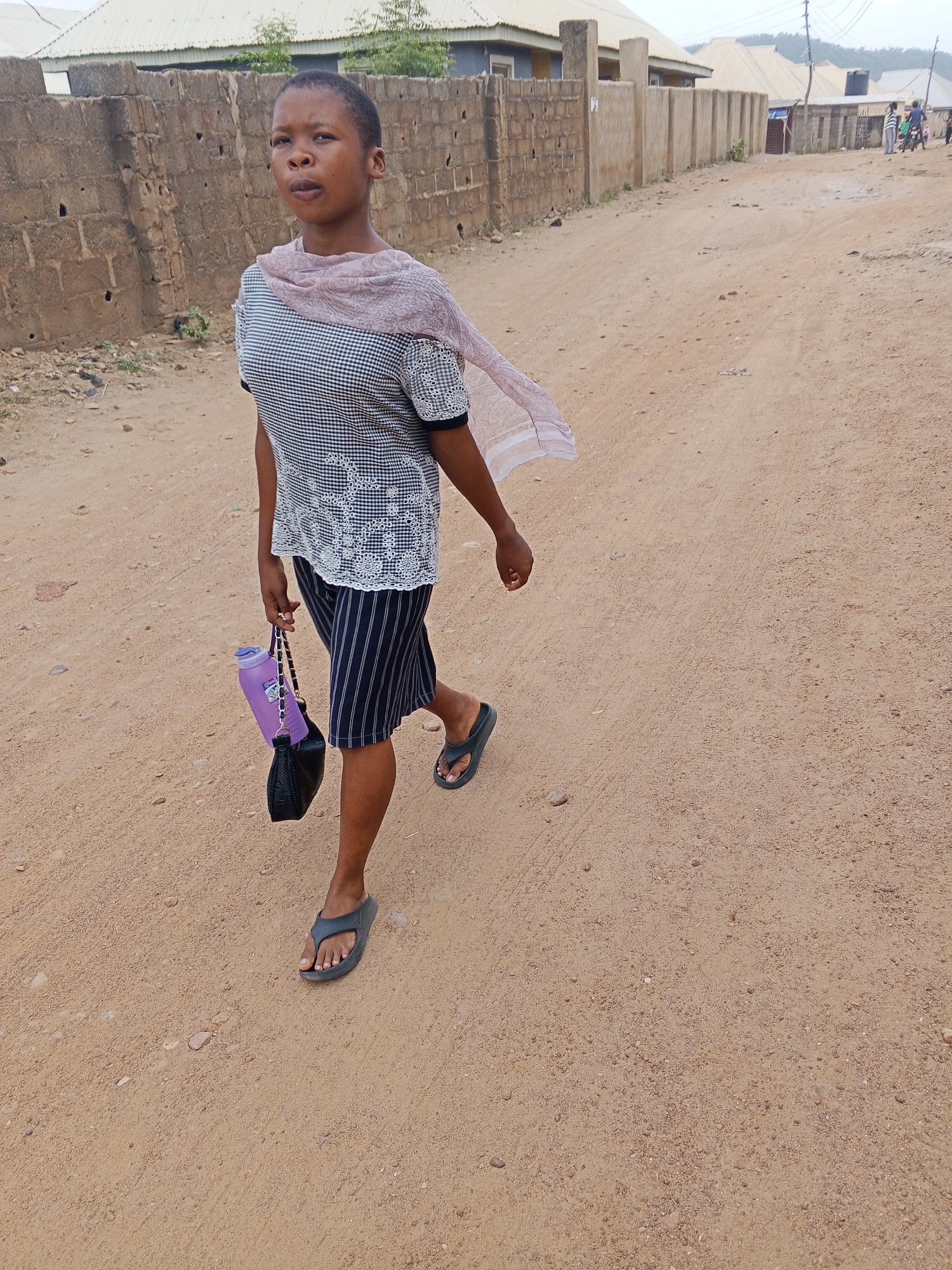 A woman walks on a dirt road in Adankolo, Nigeria. She wears a checkered shirt, striped knee-length skirt, and gray flip-flops. A pink scarf is draped around her shoulders. In her left hand, she carries a black purse and a purple water bottle attached to the purse's strap. The road is unpaved with a mixture of dirt and small rocks. Buildings and fences line both sides of the road. Other people are visible further down the road. The sky appears overcast.