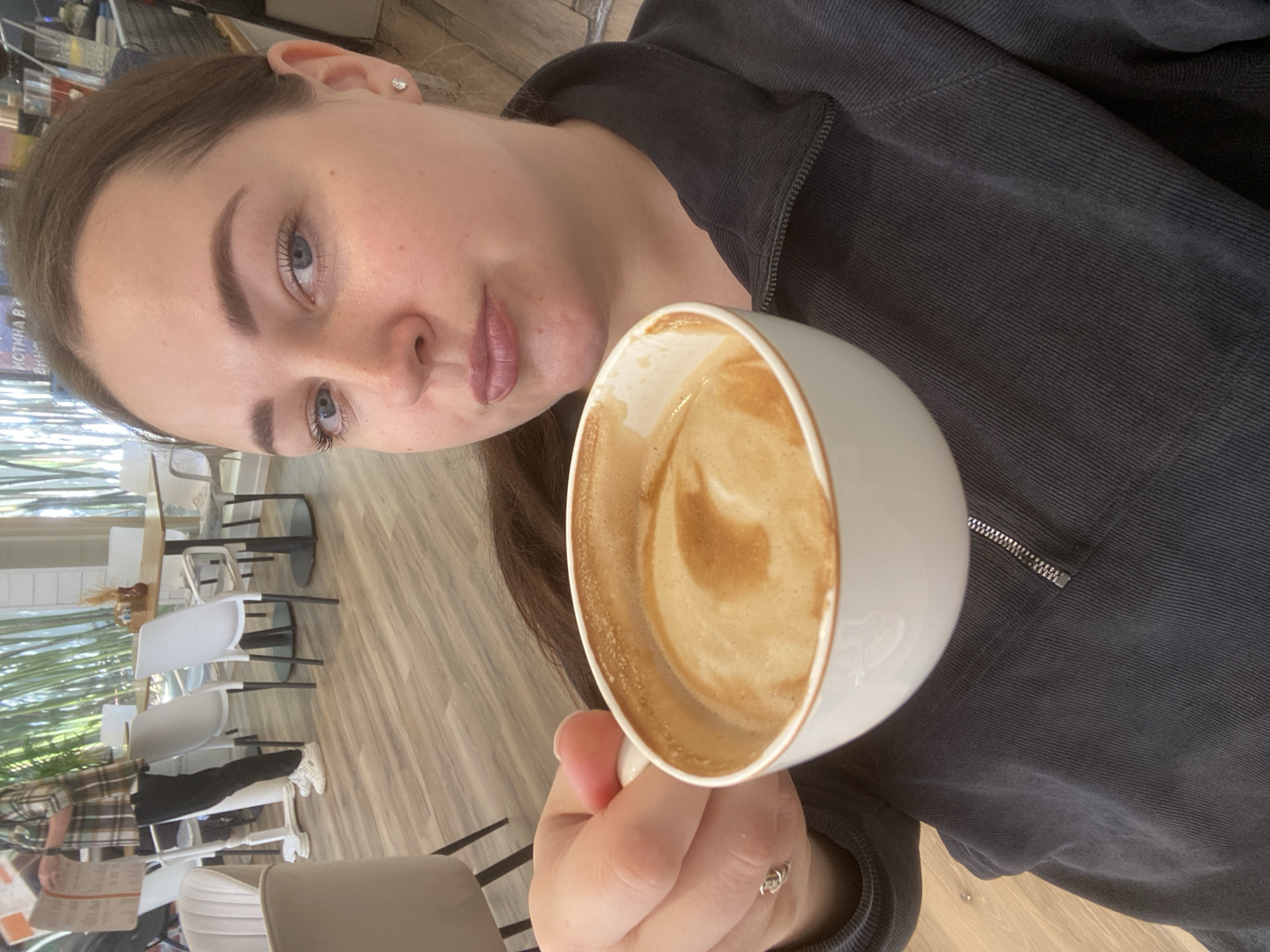 A woman with fair skin, brown hair, and blue eyes is holding a white coffee mug with latte art in her right hand. She is wearing a dark grey zip-up sweater. The background shows a brightly lit interior space, possibly a cafe, with tables and chairs arranged neatly. There are plants visible outside the window, and the overall setting suggests a modern and clean environment. The image doesn't confirm the location in Sochi, Russia.