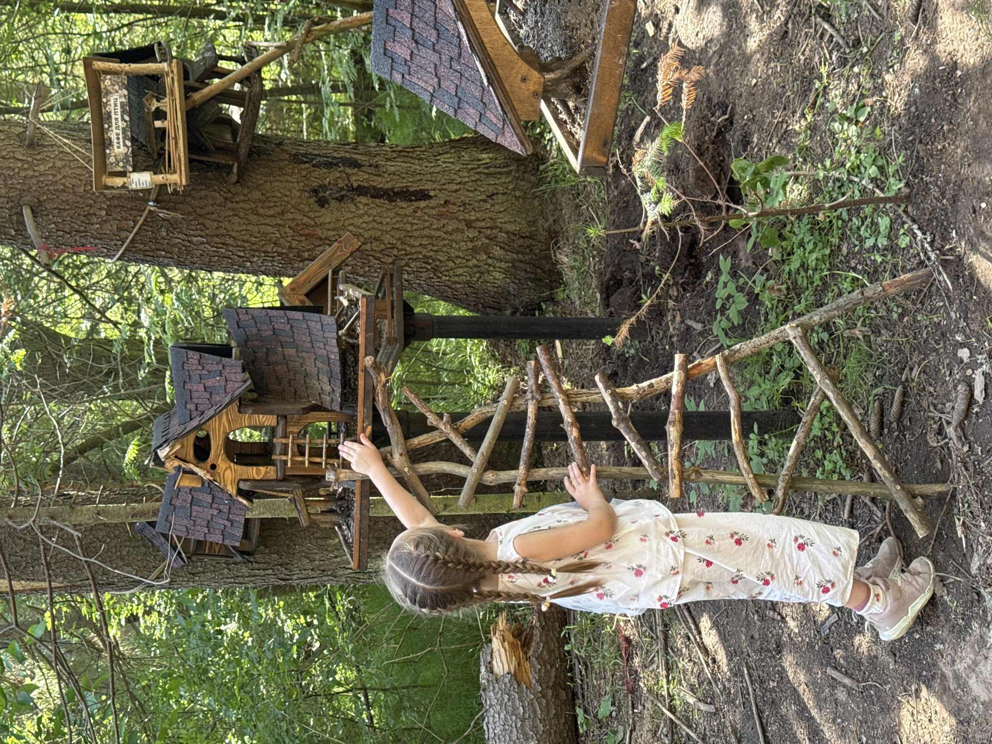 The image depicts a young girl, viewed from behind, with braided hair, standing in a wooded area and reaching towards a miniature wooden house structure attached to a tree. She is wearing a white outfit with a floral pattern and light-colored shoes. The structure resembles a birdhouse or a fairy house and is elaborate, featuring multiple levels and a ladder. In the background, other similar miniature structures are visible, also mounted on trees. The environment is a forest floor with dirt, fallen leaves, and sparse vegetation. One of the birdhouses has a sign with Russian text that translates to "Cafe for Birds." The scene is set in the city of Votkinsk, Russia, likely a park or nature area with interactive art installations.