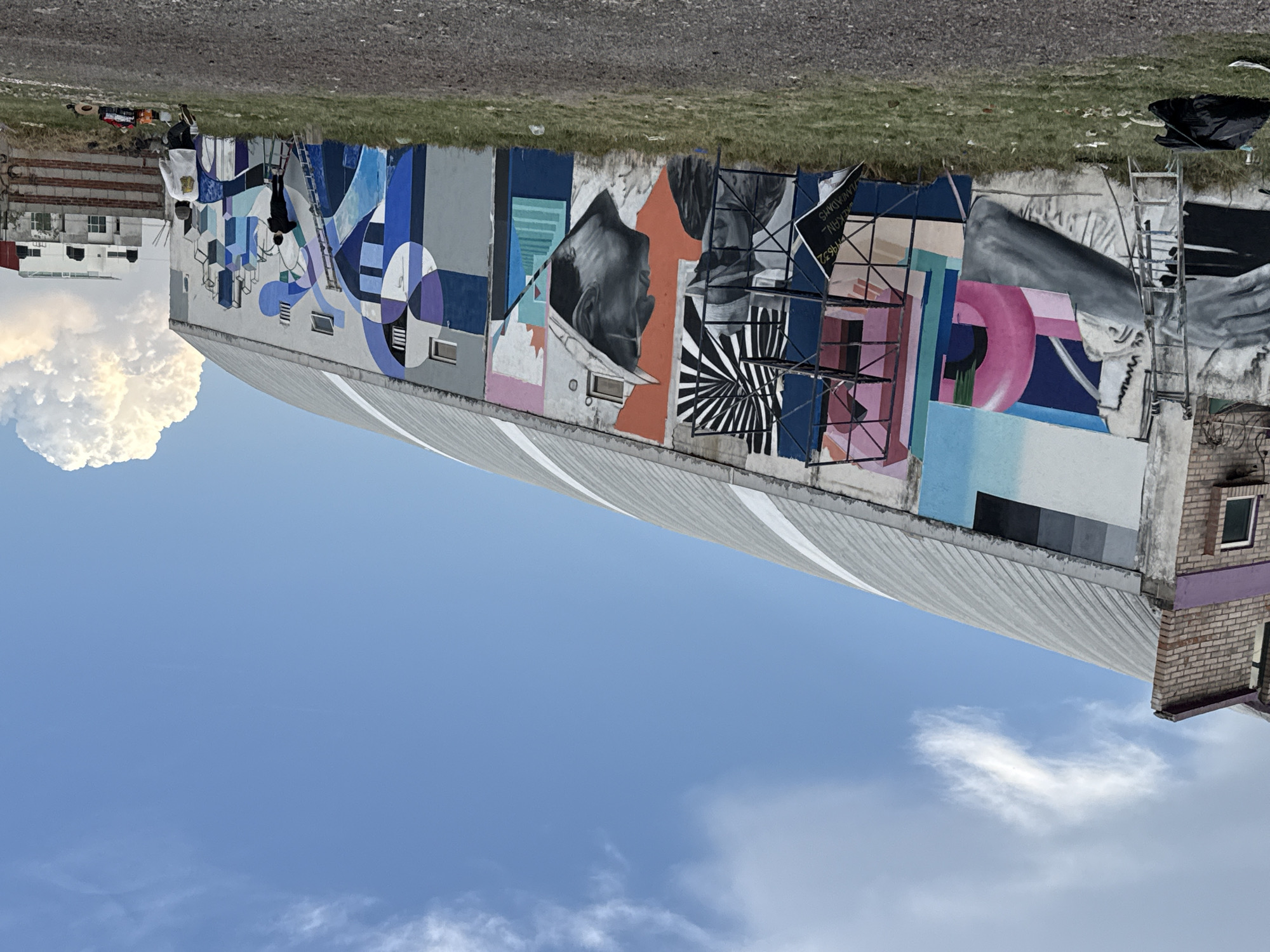 A mural on a building in San Andrés Cholula, Mexico, shows various abstract shapes and human portraits. An artist works near the left edge of the mural on scaffolding. A large cumulus cloud is visible next to the building. The sky is blue with scattered cirrus clouds. A brick structure adjoins the building on the right. The mural appears to be unfinished, with scaffolding, ladders, and signs present.