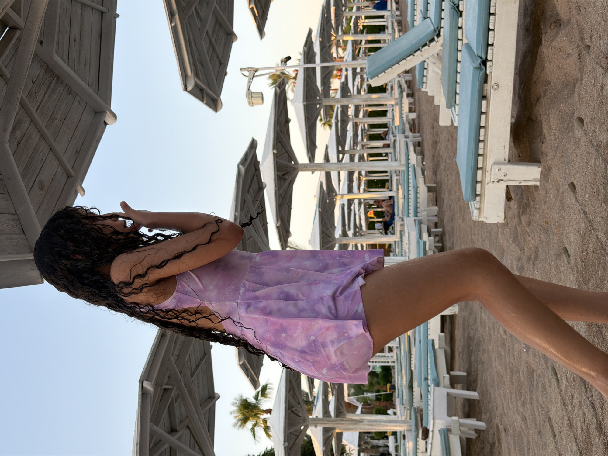 A young girl with long, dark, curly hair is standing on a sandy beach in Sharm El Sheikh, Egypt. She is wearing a light purple swimsuit with a galaxy-like pattern. Her hair is wet and appears to have been recently washed. She is looking to her right, with her right arm raised to her face as if shielding her eyes from the sun or brushing away water. The background shows a row of empty white lounge chairs with blue cushions, partially covered by thatched umbrellas, suggesting a resort or beach club. Palm trees are visible in the distance, indicating a warm climate. The lighting suggests it is late afternoon or early evening, with the sun casting a warm glow. The overall mood is relaxed and carefree, typical of a beach vacation. There is no visible text in the image.