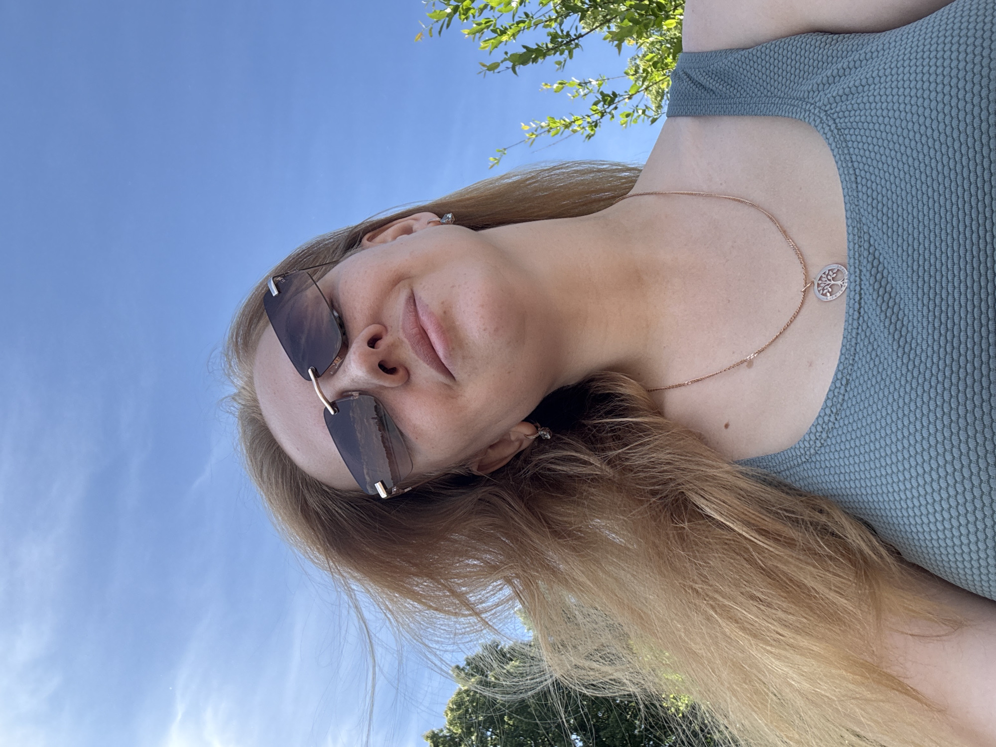 A low-angle, close-up outdoor photograph captures a woman from the chest up, posing for a selfie against a bright blue sky. She has fair skin, light brown hair, and is wearing dark sunglasses with gold accents. A delicate rose gold necklace with a tree of life pendant rests on her chest, and she wears a textured, light green tank top. Her right ear, adorned with a small hoop earring, is visible. Behind her, out-of-focus green foliage and trees are visible to the left and right. The sky is clear with a few wispy white clouds. The location is unknown, and the time of day is likely daytime due to the bright lighting and clear sky.
