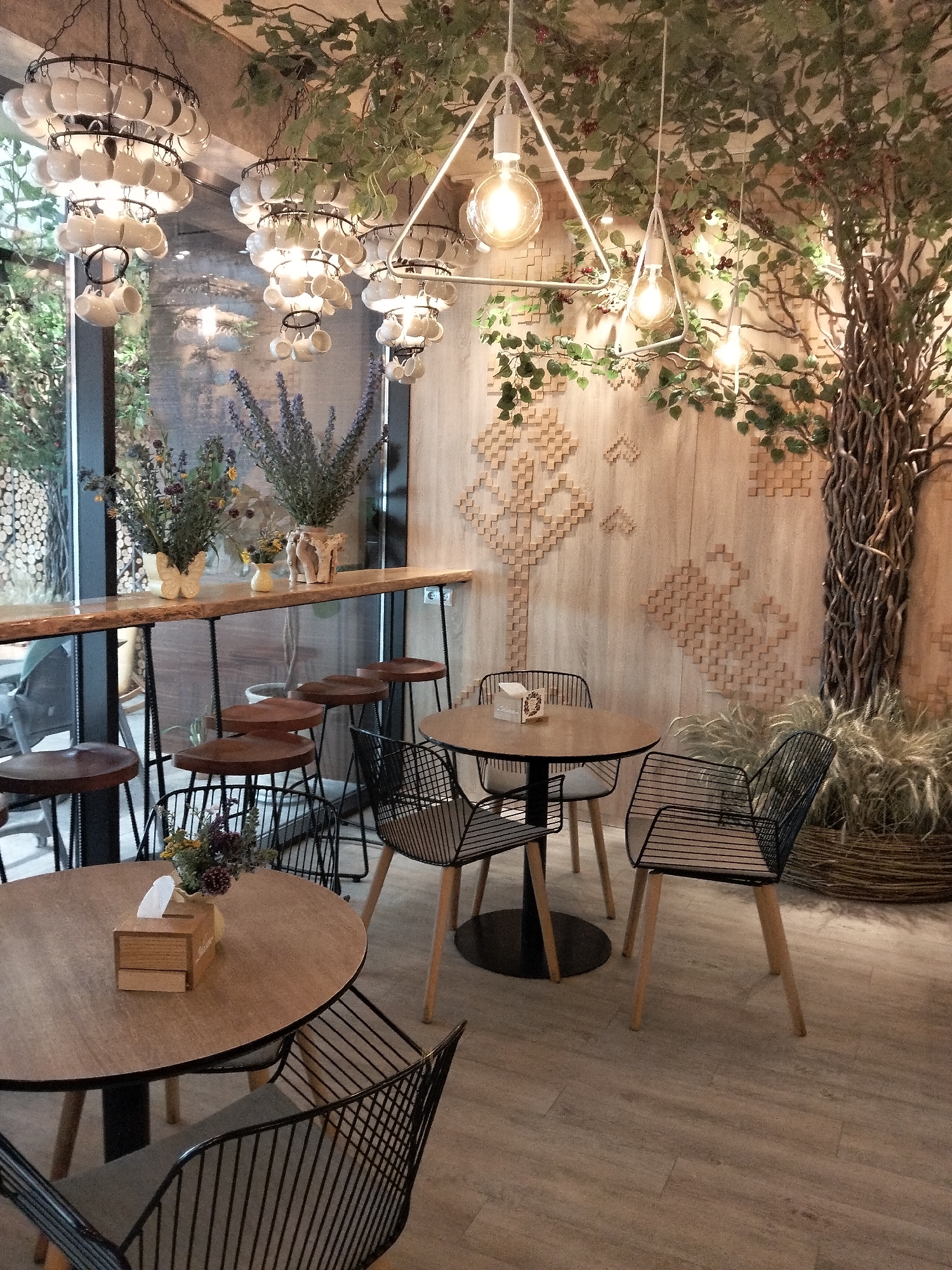 The image depicts the interior of a cafe in Peresecina, Moldova. The cafe features a rustic and natural aesthetic, with a large, artificial tree with branches and leaves creating a canopy overhead, interspersed with decorative lighting fixtures. The walls are decorated with a textured, geometric pattern resembling a tree or abstract design, and a large, faux tree trunk is positioned in the corner.
Several tables and seating arrangements are visible. A long, wooden bar counter runs along the back, equipped with several wooden stools with black metal bases. In the foreground, two round wooden tables with black wireframe chairs are set up. One table has a small wooden tissue box with a white tissue peeking out, and a small vase of dried flowers. The floor is made of light-colored wooden planks.
The overall ambiance suggests a cozy and nature-inspired dining or coffee space. There are no people visible in the image. The lighting appears to be a mix of ambient and decorative, with exposed bulbs and light filtering in from what seems to be a window or glass partition in the background, revealing a glimpse of an outdoor area with more trees.