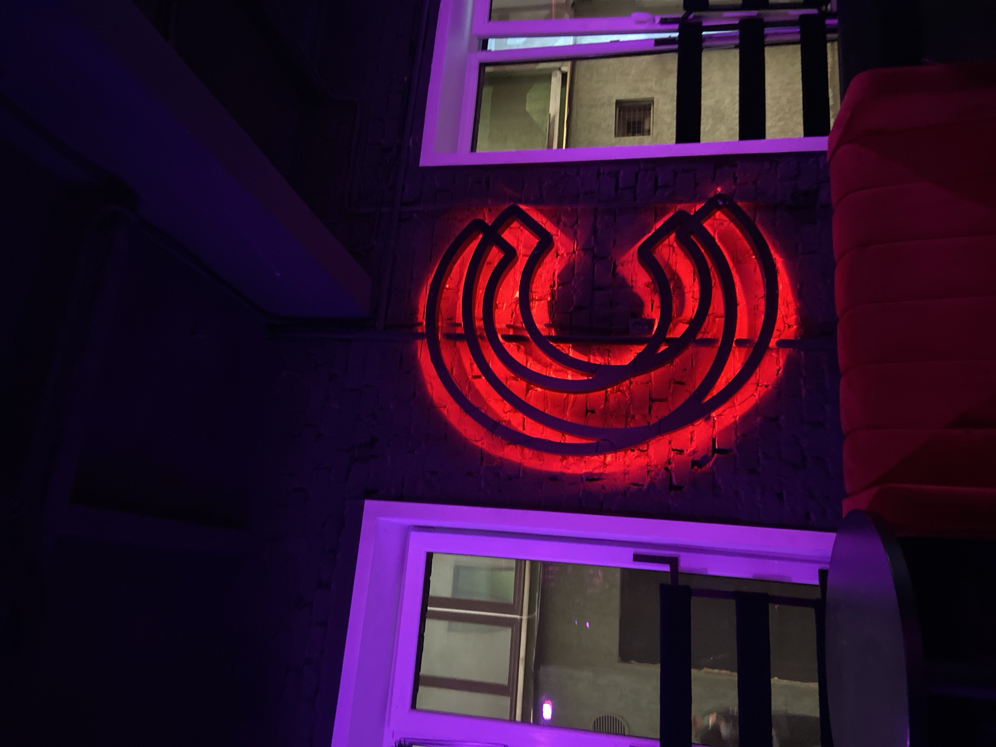 The image depicts the interior of a dimly lit establishment, possibly a bar or lounge, featuring purple ambient lighting and a prominent red neon sign shaped like a stylized "C" on a brick wall. To the left and right of the sign are closed doors or windows with white frames, illuminated by the purple light. In the foreground, a red upholstered sofa is visible, and to the left, a dark circular table and a black chair are present. The overall atmosphere suggests a modern, possibly trendy, urban setting. The location in Brest, Belarus, is not discernible from the visual information alone.