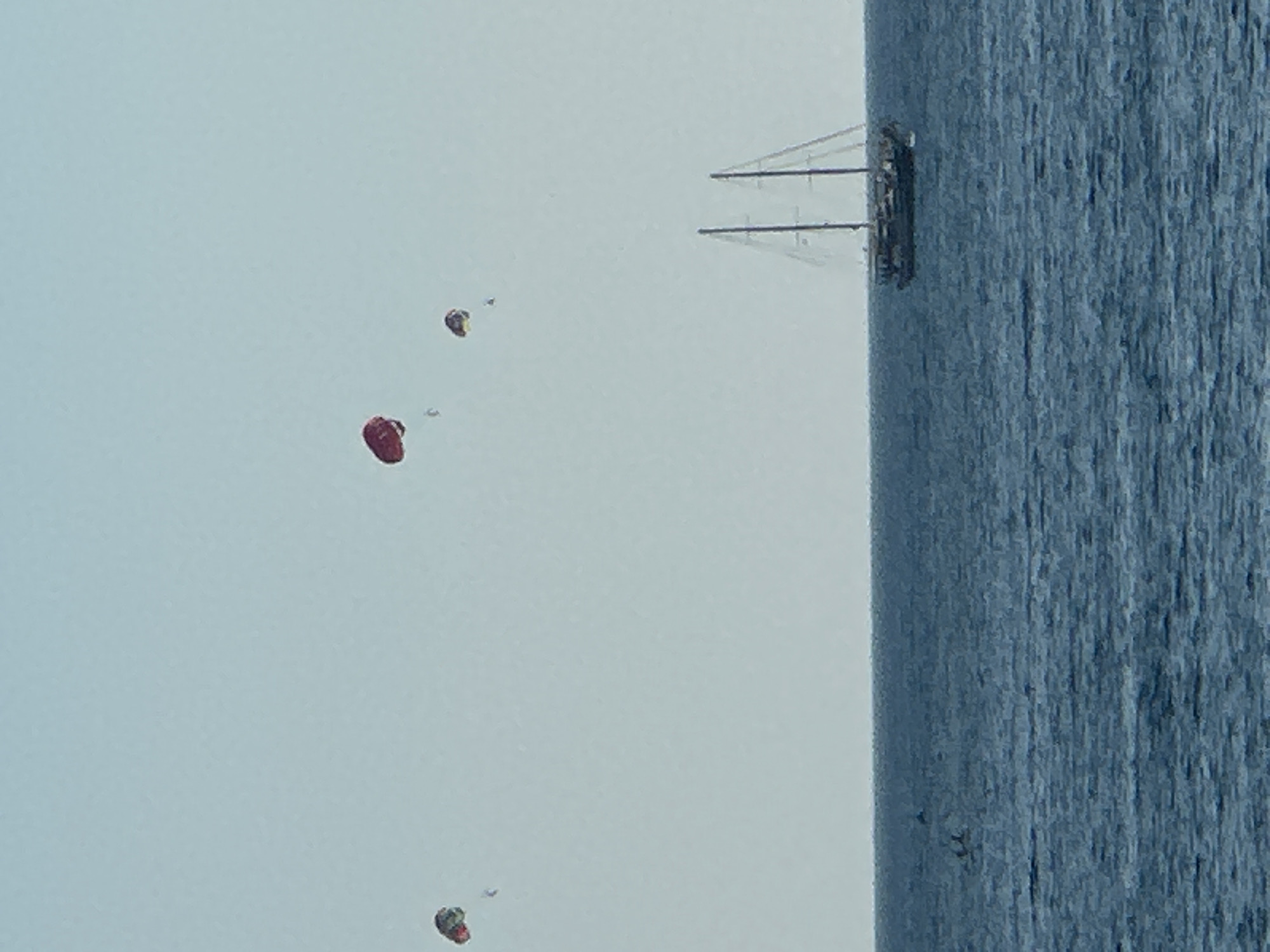 The image captures a serene day at sea, likely off the coast of Budva, Montenegro, given the presence of parasailing and a traditional sailing vessel. In the distance, three colorful parachutes are visible against a clear, pale blue sky, indicating people are engaged in parasailing. One prominent red parachute is in the upper-middle of the frame, with two smaller parachutes to its left and right, respectively.
Dominating the lower right portion of the image is a magnificent sailing ship with two masts. The ship appears to be a traditional schooner, with its sails furled and its hull dark against the deep blue sea. A few individuals can be seen on the deck of the ship. The sea itself is a calm, deep blue, with subtle ripples on its surface. In the far left of the image, a small dark shape in the water might be a distant boat or a sea creature. The horizon line is clearly defined, separating the sea from the sky, which has a slight gradient of color. The overall impression is one of leisure and outdoor activity on a bright, clear day.