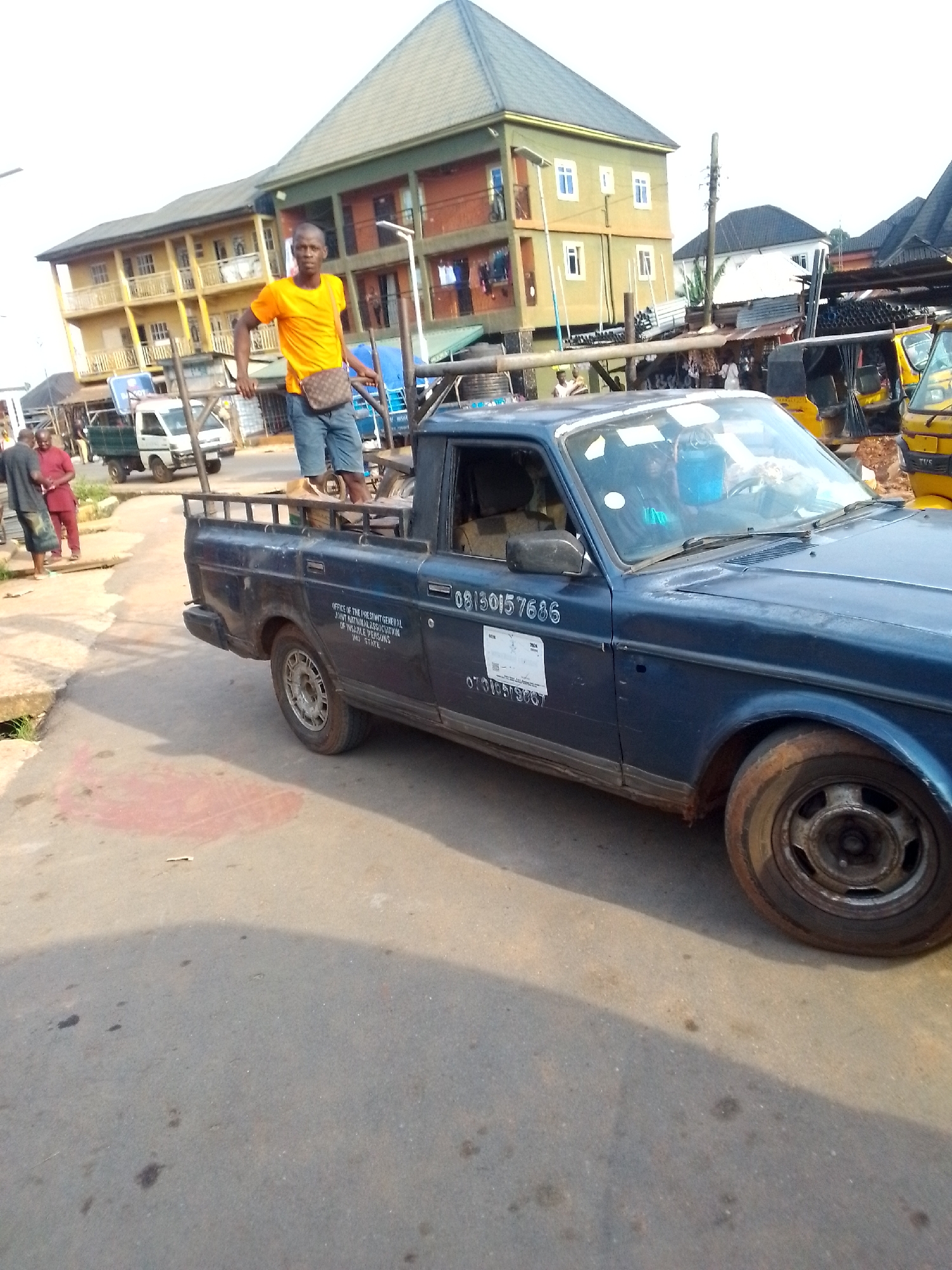 The image depicts a scene in Orlu, Nigeria. A blue pickup truck is parked on a paved street. A man in an orange t-shirt and denim shorts is standing on the truck bed, holding onto the truck's roof rack. He also wears a brown bag across his body. A building with multiple stories and a green roof can be seen in the background. There are other vehicles and pedestrians present in the vicinity. The street pavement shows signs of wear and tear. The truck has the text "OFFICE OF THE PRESTANT GENERAL JOINT NATUMALASSOCIATION OF INSABLE PERSONS IMO STATE" and a phone number on its side.