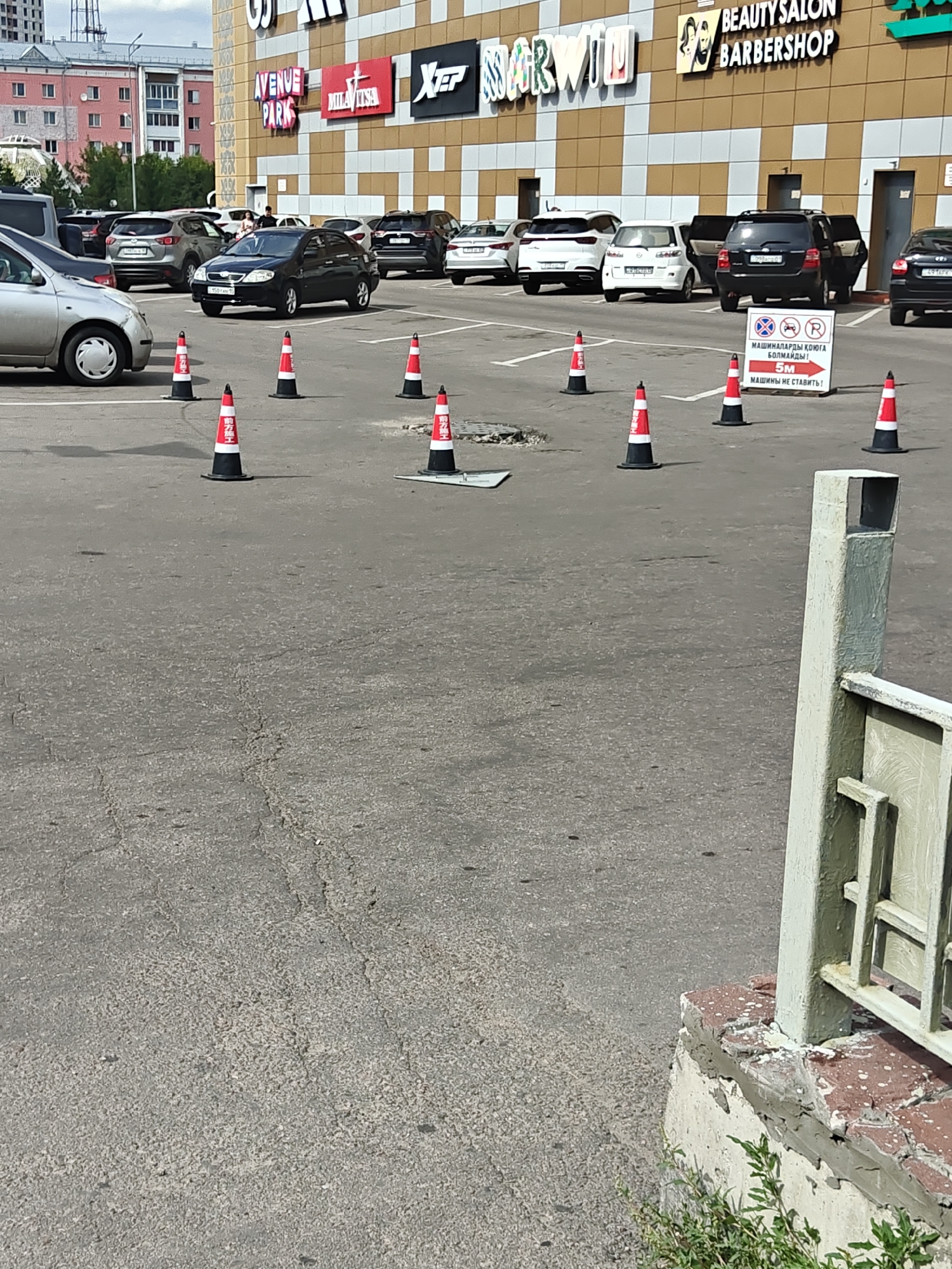 The image shows an outdoor asphalt parking lot in front of a large building with a textured facade, possibly a shopping center or a business complex, in Petropavl, Kazakhstan. The building has signage that includes "AVENUE PARK", "MILLATISA", and "BEAUTYSALON BARBERSHOP". Several cars are parked in the lot, including a black sedan, a black SUV with its doors open, and several white SUVs. The foreground is dominated by the asphalt surface, which has visible cracks and is marked with several orange and white traffic cones. In the middle ground, a white sign with red text and symbols is posted near the black SUV. The sign appears to have a prohibition symbol and text in what seems to be Cyrillic and possibly Latin script, indicating parking restrictions. A light-colored metal post and railing are visible in the far right of the image, partially obscuring the view of the parking lot. The sky is visible in the upper portion of the image and appears to be overcast with some blue sky peeking through. The overall scene depicts a typical urban parking area on a daytime.