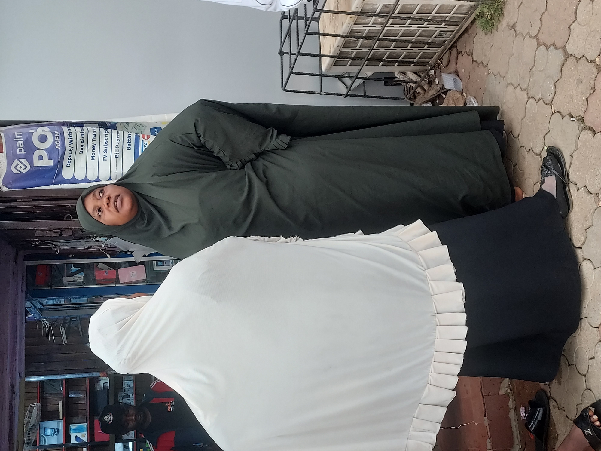 The image captures a daytime street scene in Jos, Nigeria, featuring two women in front of what appears to be a local business stall or agency. In the foreground, a woman is seen from the back, wearing a cream-colored garment with pleated details and a black skirt or dress. Beside her, a woman in a dark green abaya and hijab looks upwards with a composed expression.
Behind the women, a blue banner for a 'Palm Pay Agency' (identified by "paln PO AGEN" in the OCR) is prominently displayed. The banner lists various services offered, including "Deposit/Withdrawal," "Buy Airtime," "Money Transfer," "TV Subscription," "Bill Payment," and "Betting." Inside the open-fronted stall, shelves are stocked with what appear to be electronics and other goods, with a man's head partially visible on the left, wearing a cap. The ground is paved with interlocking concrete blocks, and the feet of several individuals are visible at the bottom right, wearing various footwear. A black metal rack is situated at the top right of the frame. The bright lighting indicates it is daytime. The overall setting suggests a common commercial or market area in the city.