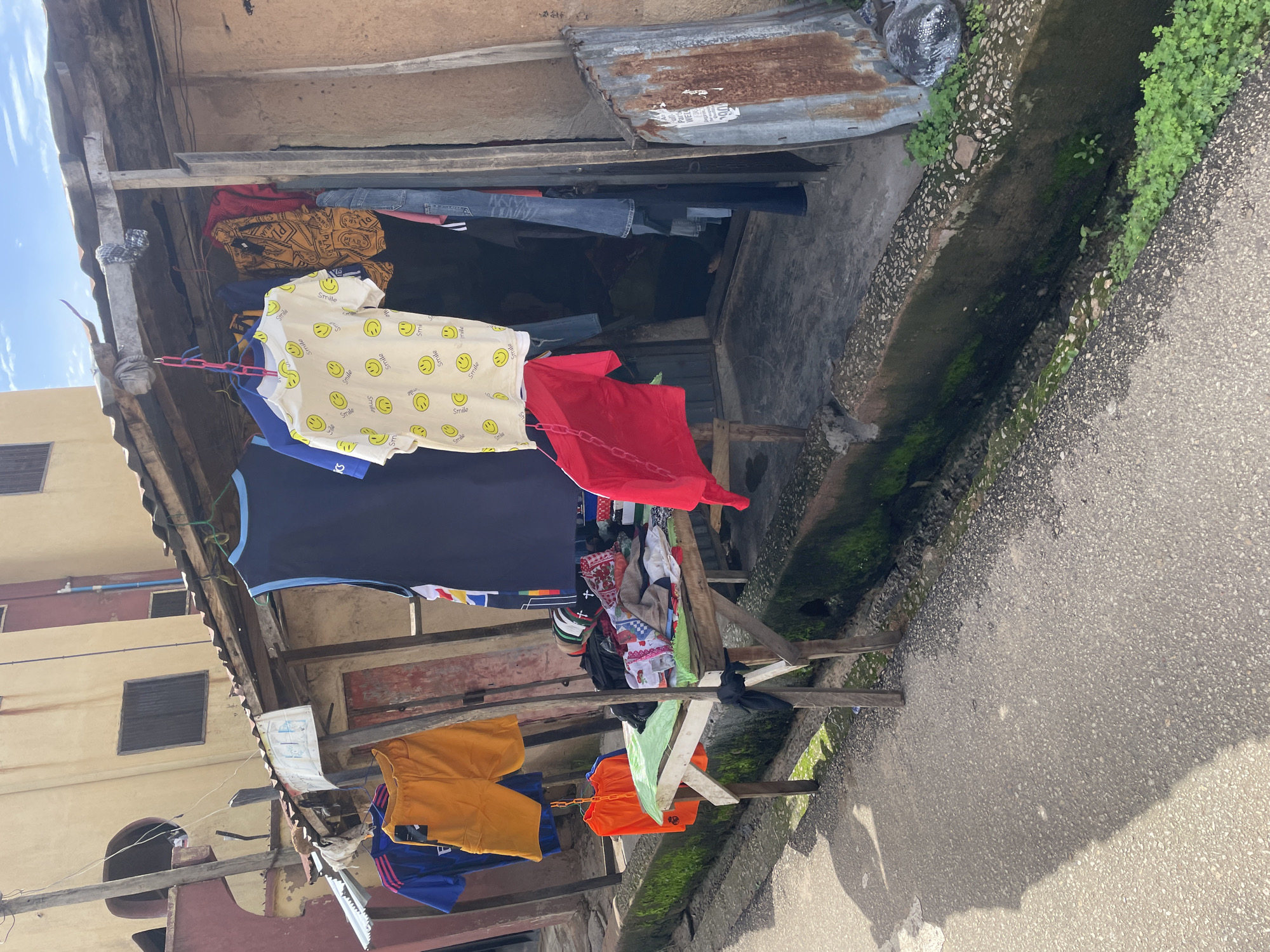 The image captures a street-level view of an outdoor market stall in Jos, Nigeria. The stall appears to be a temporary structure, with a corrugated metal roof supported by wooden posts. Various clothing items are displayed, hanging on lines strung across the front of the stall. These include t-shirts with smiley face patterns, athletic wear, shorts, and what appear to be jeans. Some clothing is also piled on a wooden table in front of the stall. The ground surface is a mixture of asphalt and a concrete drainage channel lined with moss and small plants. In the background, a multi-story building with yellow and red facades is visible, suggesting an urban or semi-urban setting. The overall impression is one of a vibrant, informal commercial space.