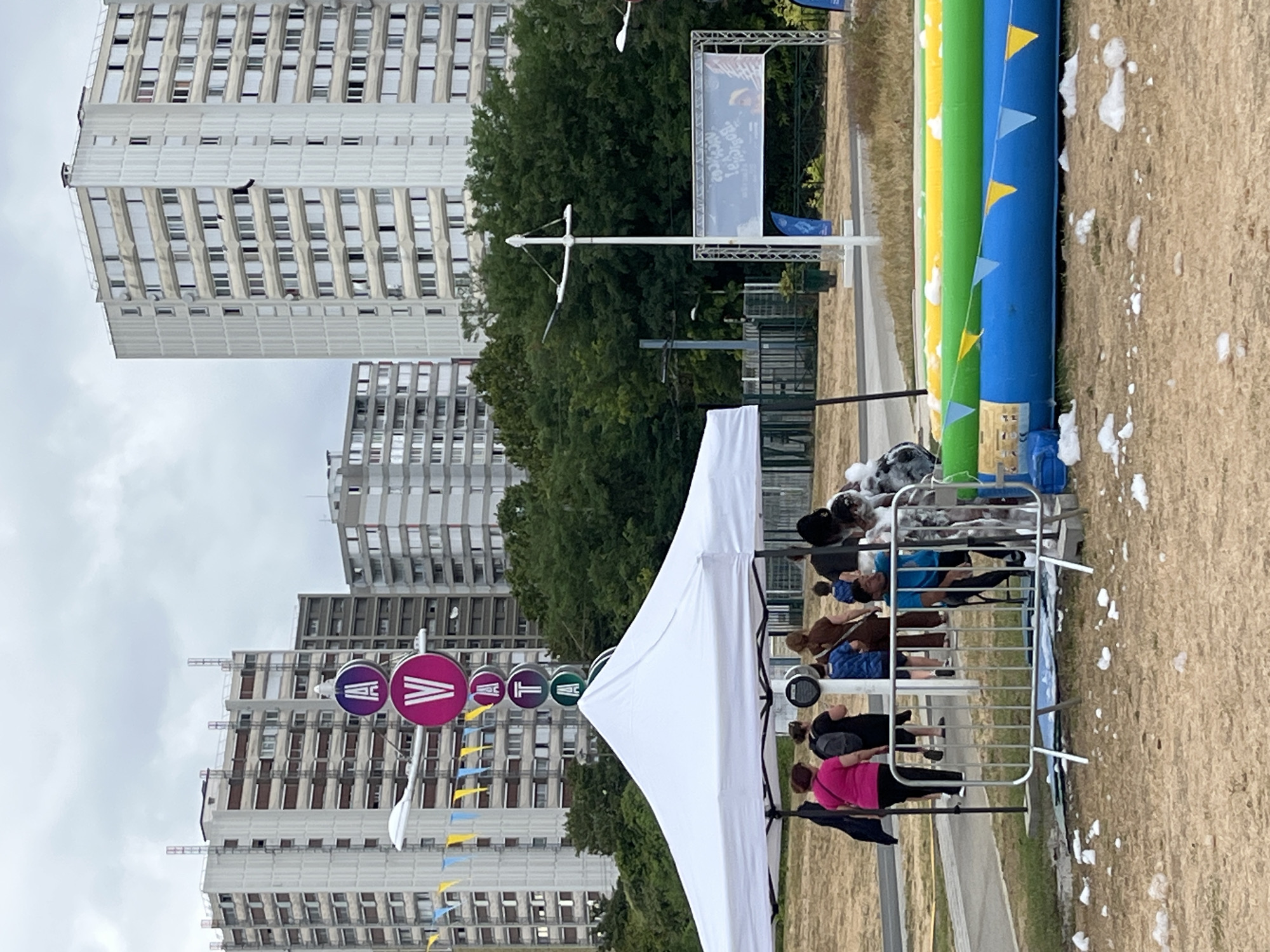 The image depicts an outdoor event in Bobigny, France, with tall residential buildings in the background. In the foreground, a white tent is set up, and a group of people are gathered around it. Several individuals are seen interacting, with some standing and others appearing to be in conversation. Nearby, an inflatable structure, possibly a water slide or pool, is visible, decorated with blue, green, and yellow sections and small triangular flags. The ground is covered in dry grass and scattered white foam. A metal barrier is also present, separating the event area from the surrounding ground. A sign with circular elements bearing letters "A", "V", "A", "T", and "A" is prominently displayed on a pole. The sky is overcast, suggesting a cloudy day. The overall scene suggests a public gathering or festival taking place in a park or open urban space in Bobigny.