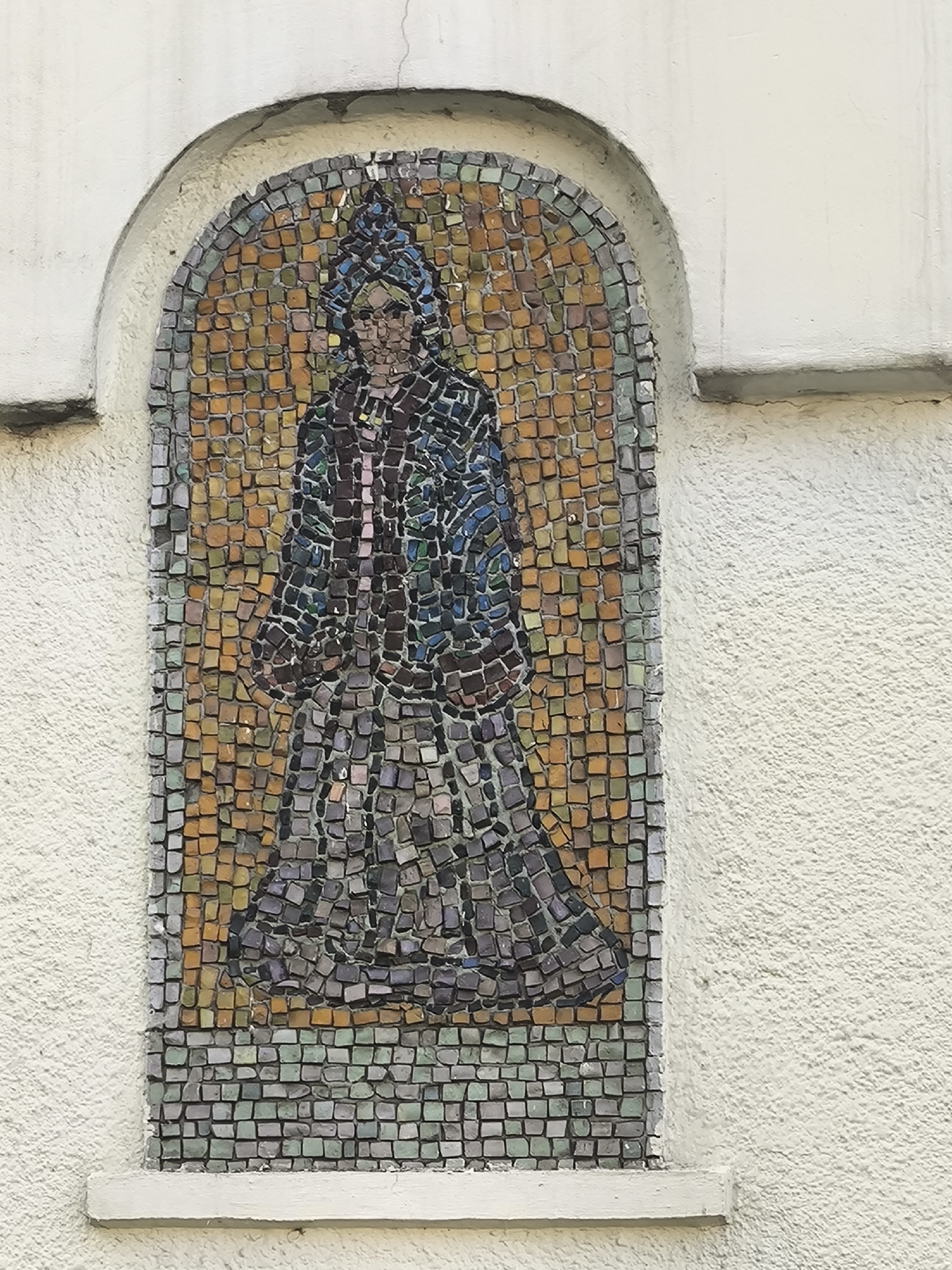 A mosaic of a person in a long robe and head covering is set into a textured, off-white wall. The mosaic is housed within an arched recess. The figure is rendered in a variety of small, colored tiles, with a predominantly purple and blue robe over a grey skirt. The background of the mosaic is composed of orange and yellow tiles. The wall surrounding the mosaic appears to be plaster, and there is a visible crack in the plaster above the arch. A narrow ledge runs horizontally below the mosaic.